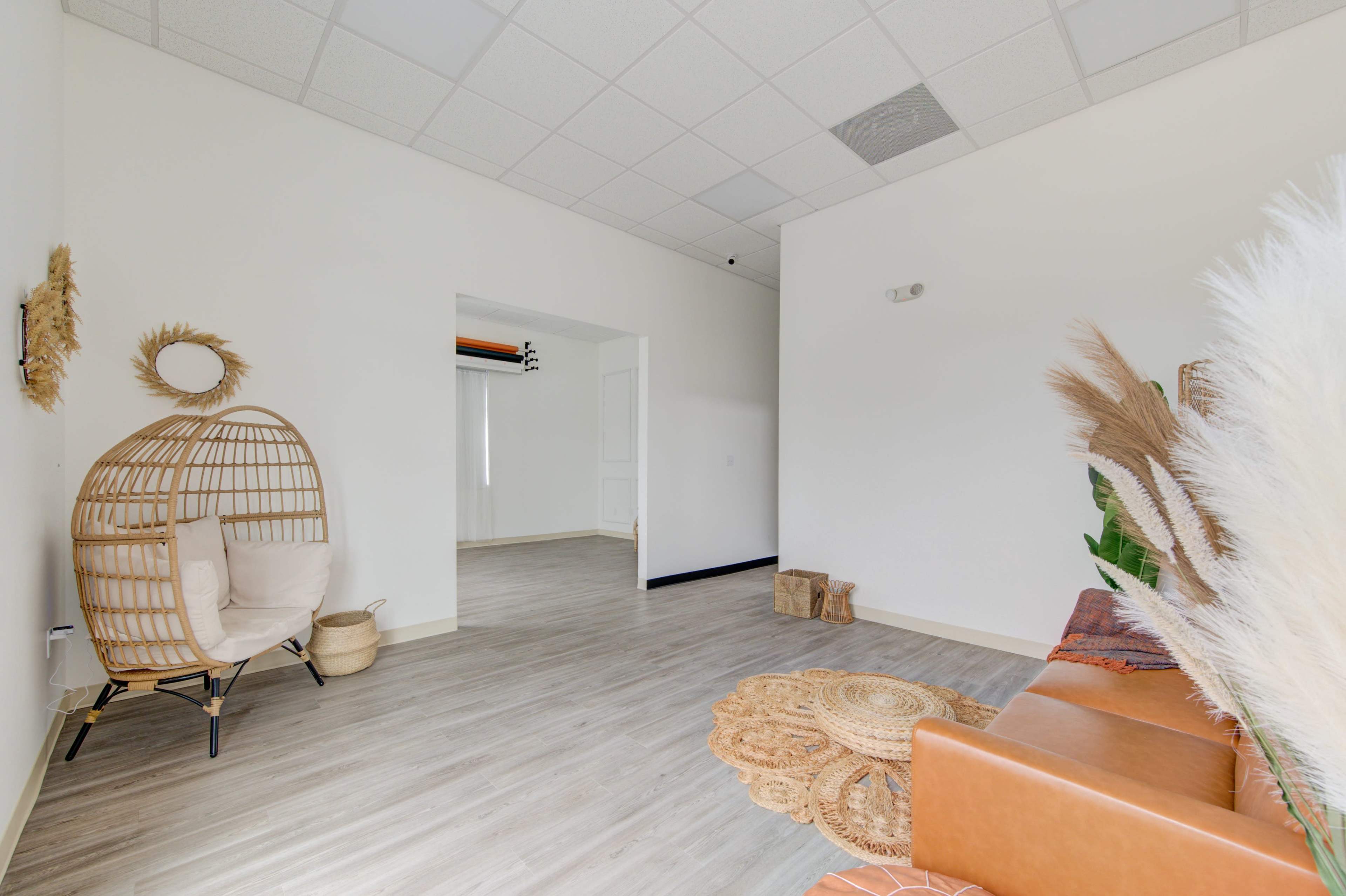 The image shows a minimalist room with a wicker chair, a brown leather sofa, and decorative natural elements placed on the floor.