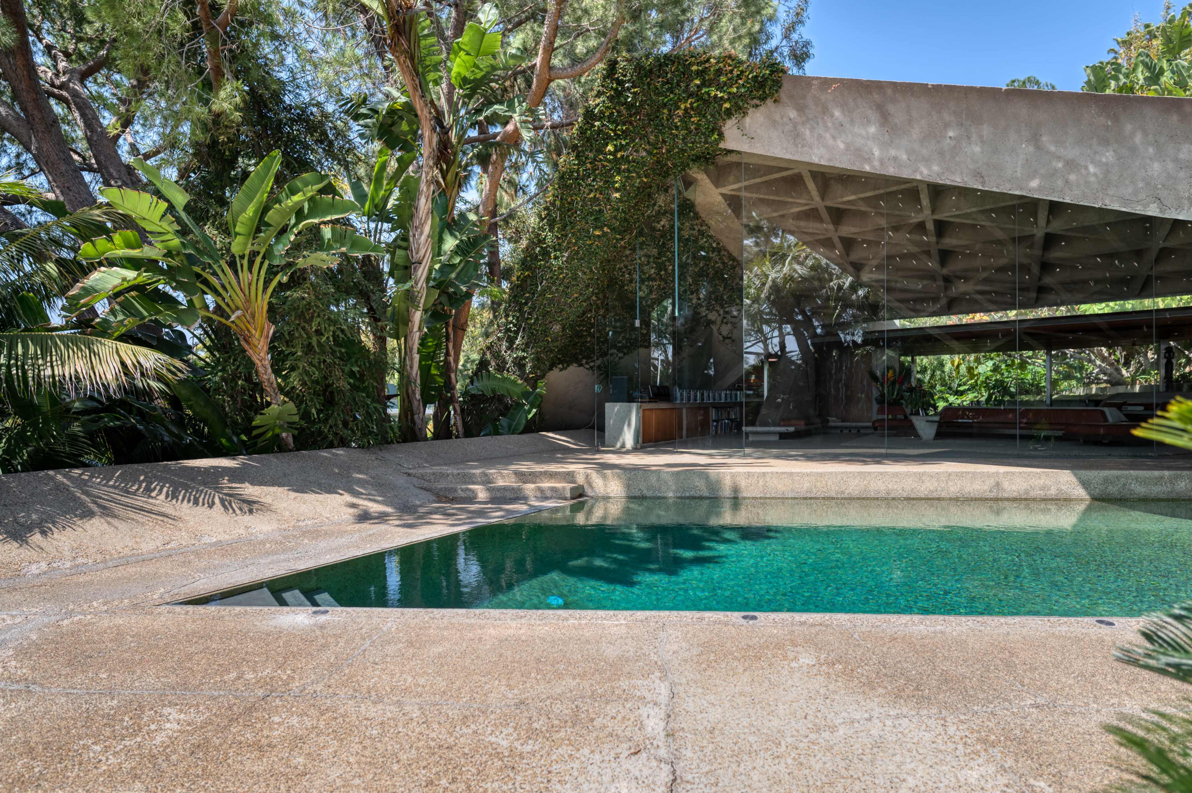 The image shows a modern architectural structure with a swimming pool surrounded by tropical plants and trees.