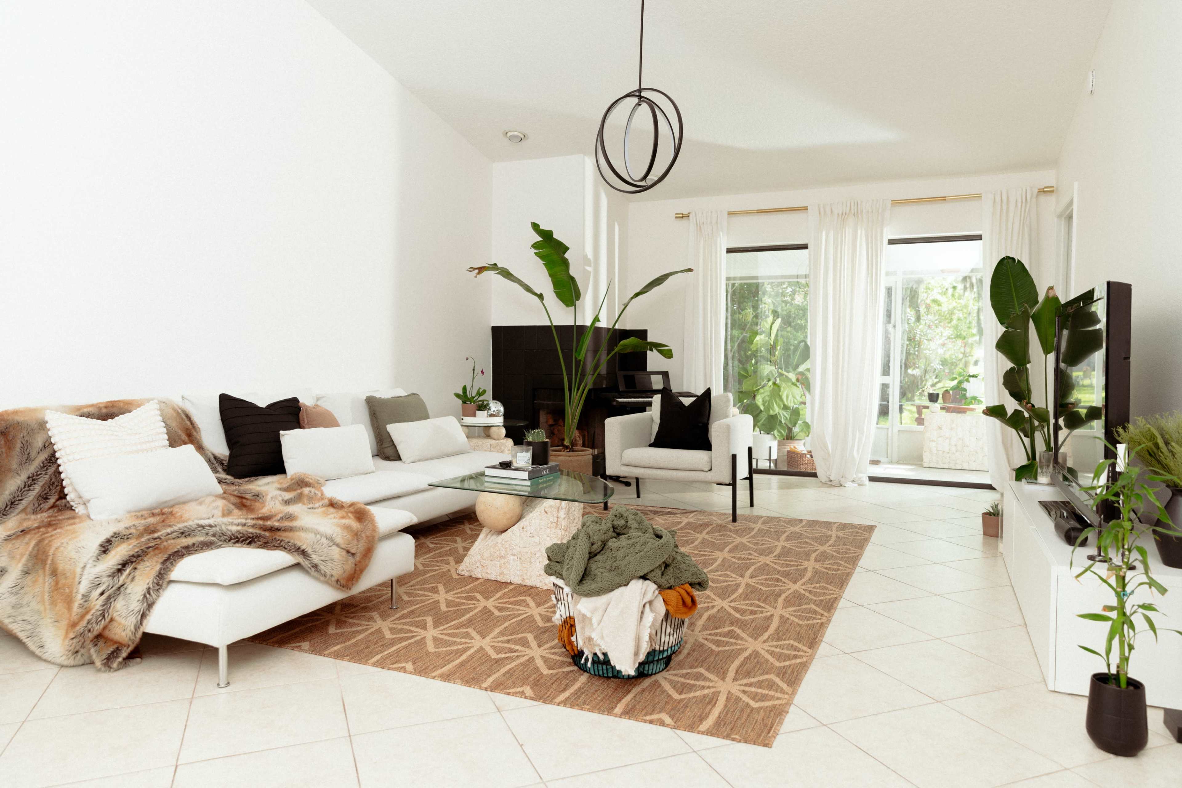 The living room features a white sectional sofa with a patterned rug, a coffee table, and large windows that let in natural light, surrounded by indoor plants.