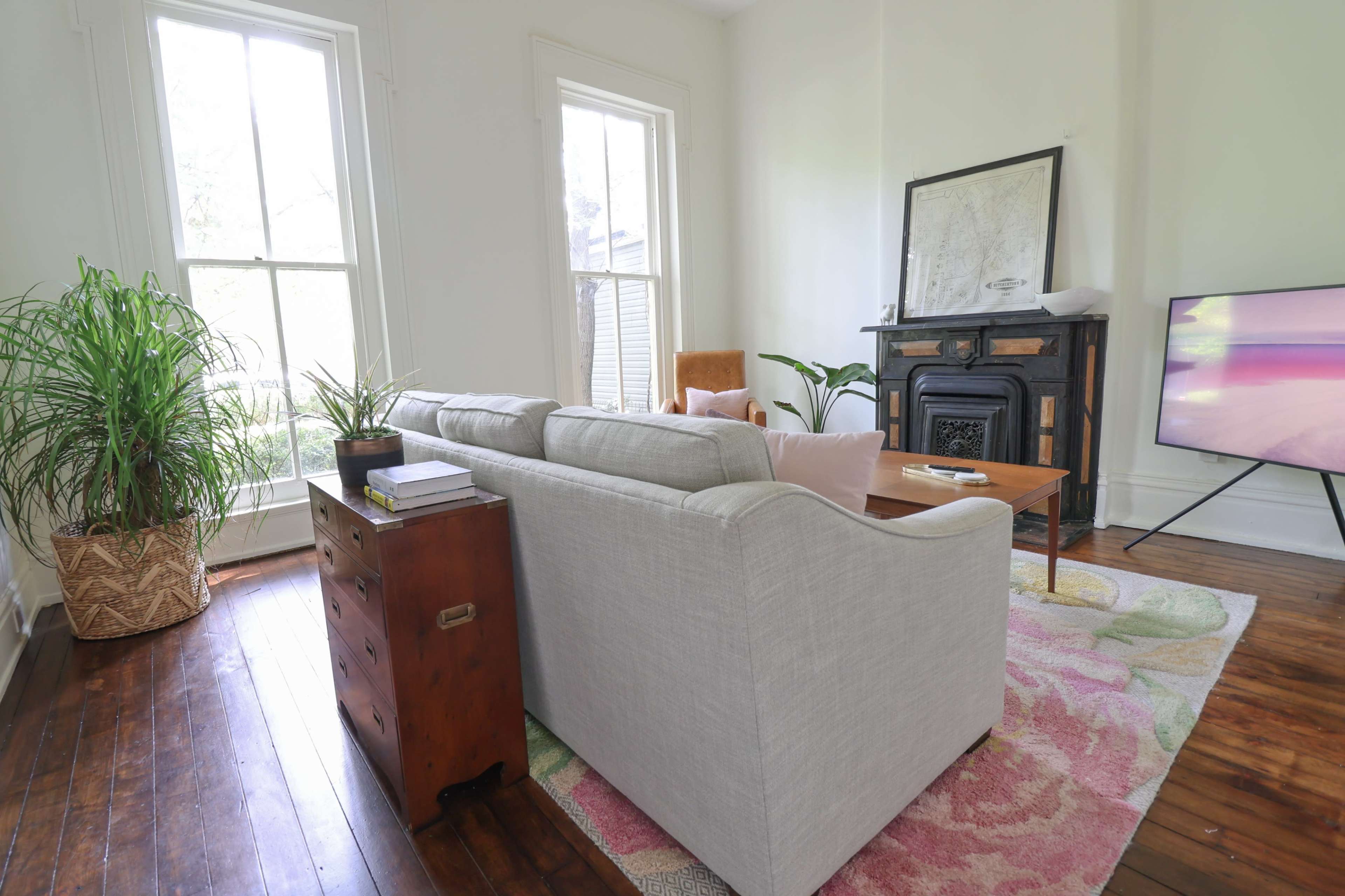 A cozy living room features a light-colored sofa, a wooden coffee table, a vintage fireplace, and a large television, with a tall plant beside a window that lets in natural light.