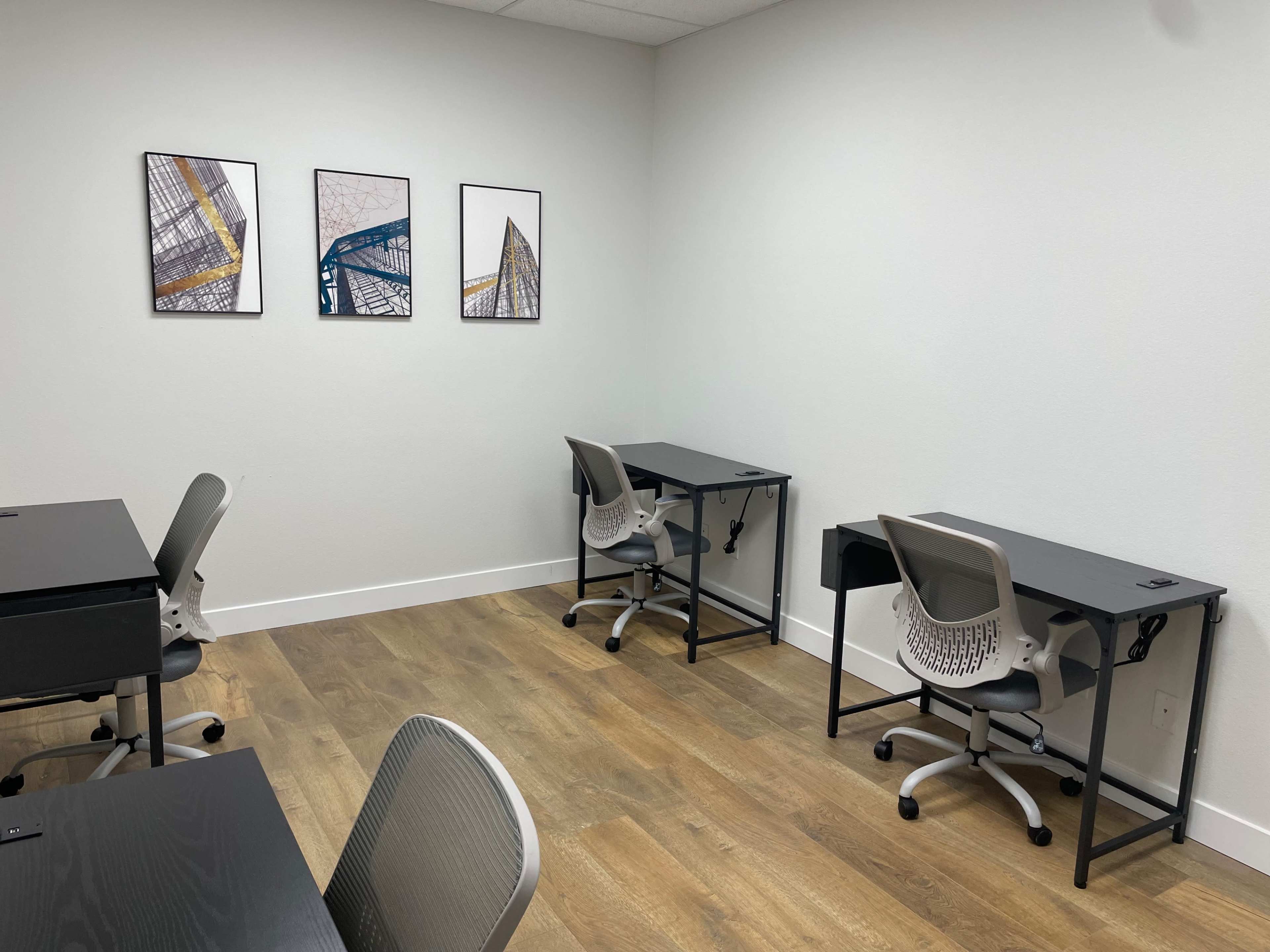 A minimalistic office space with three desks, each equipped with a chair, and three framed abstract artworks on the wall.
