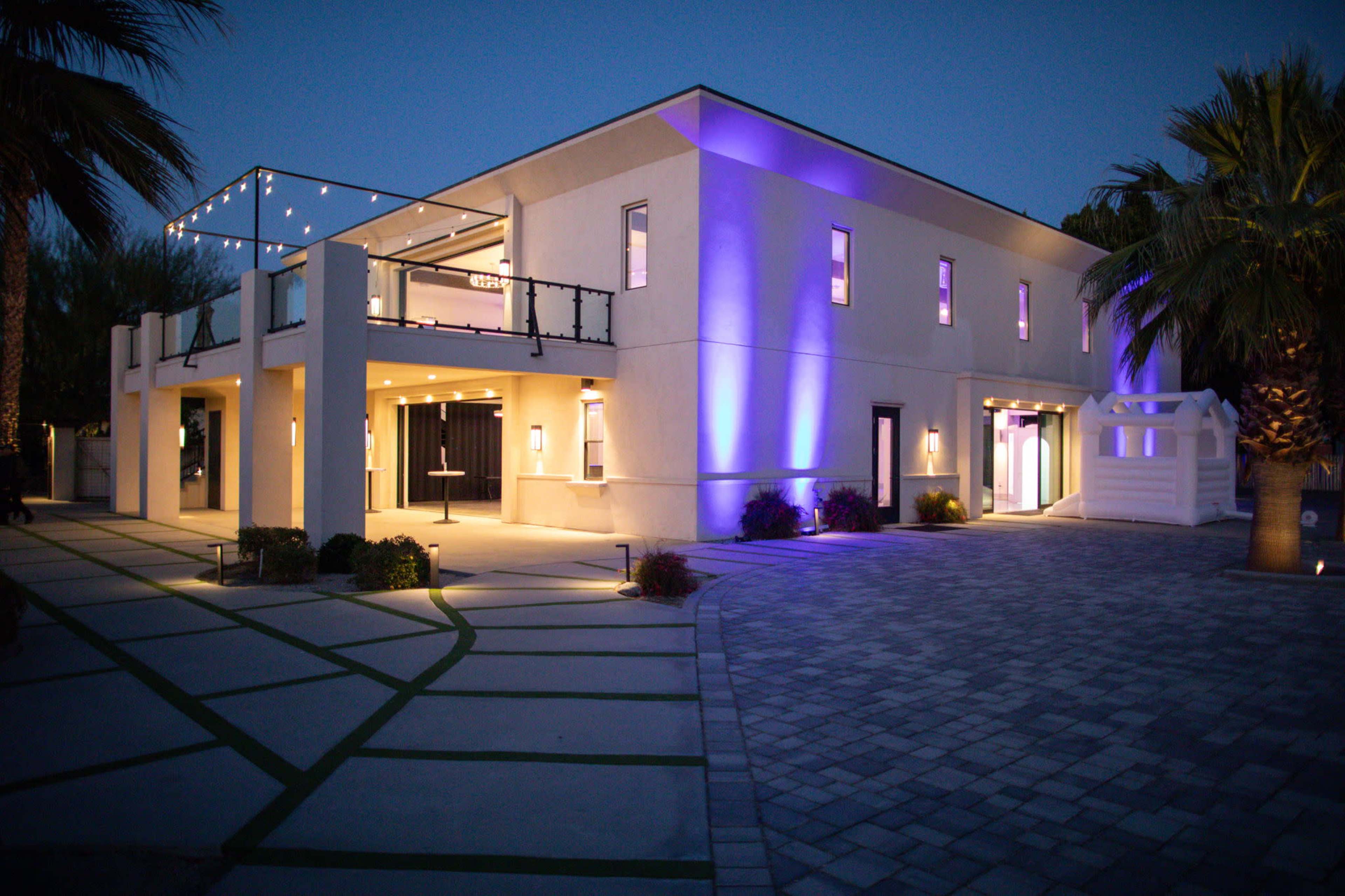 A modern building is illuminated with purple lights, featuring a spacious entrance and a patterned stone driveway under a twilight sky.