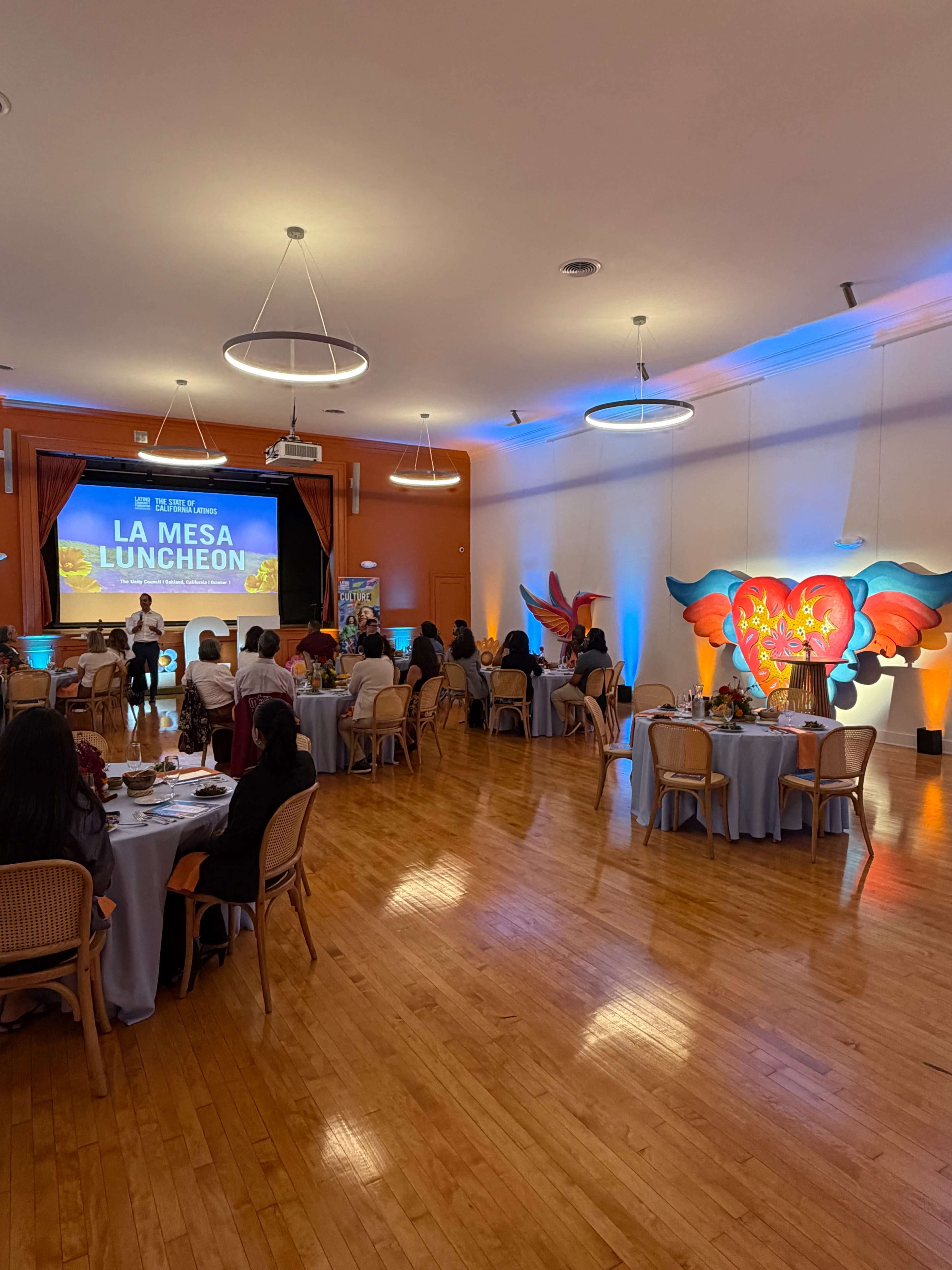 The image shows a well-lit indoor venue with guests seated at round tables, attending a luncheon event featuring a large screen displaying "LA MESA LUNCHEON."
