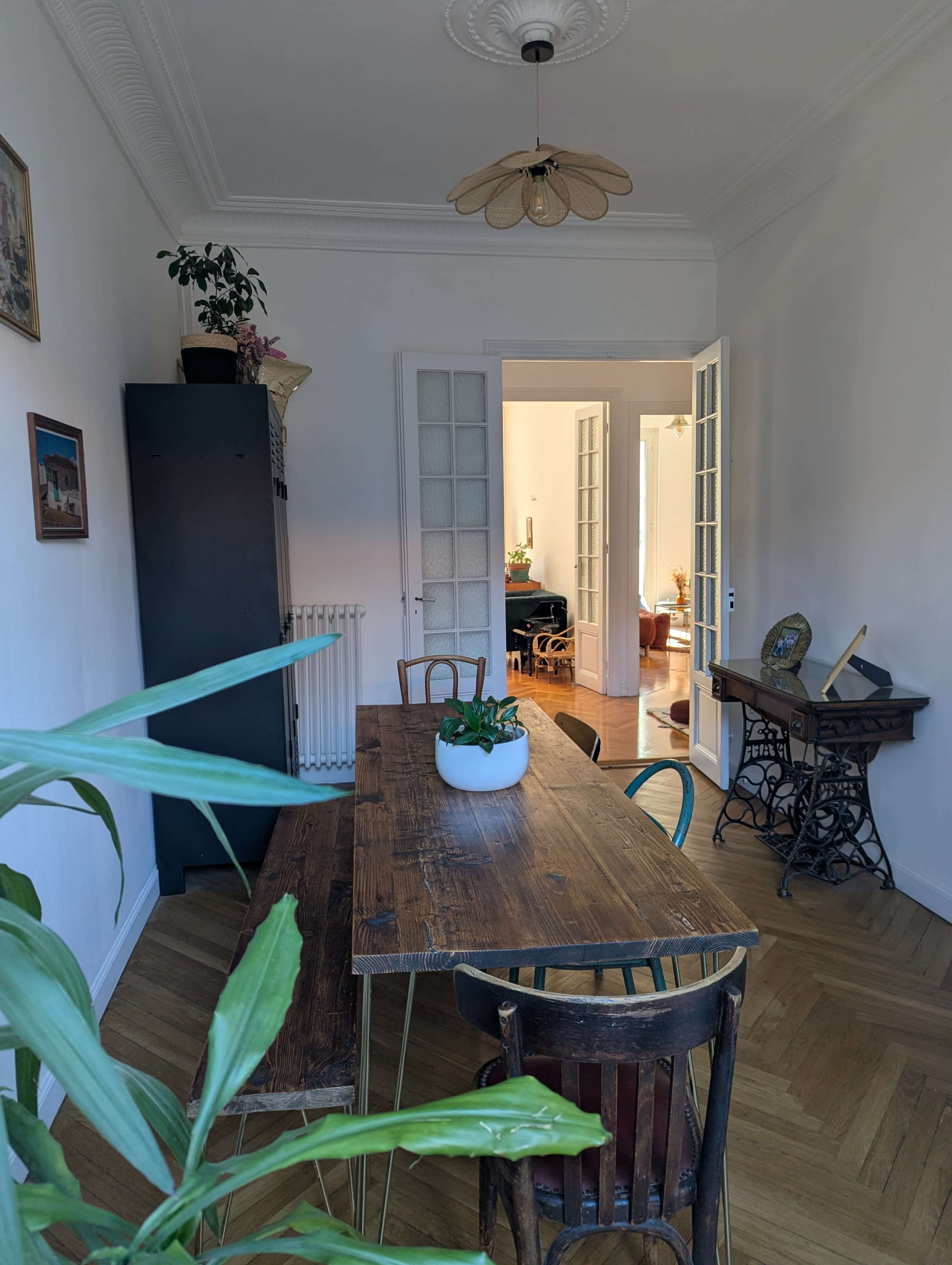 A wooden dining table with chairs is surrounded by plants in a well-lit room that leads to another room with visible furniture.