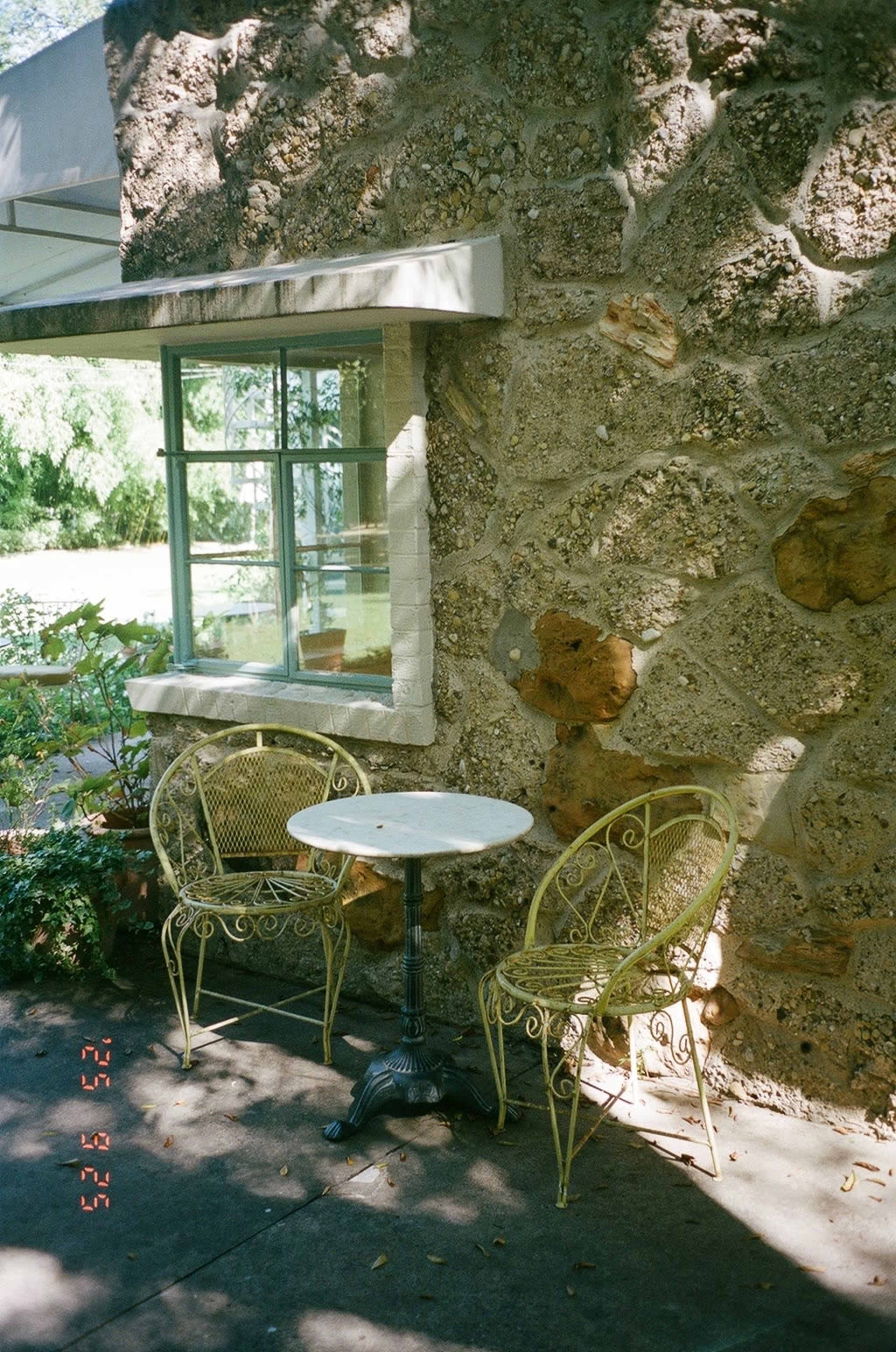 A small round table with two wrought iron chairs is positioned against a textured stone wall next to a large window.