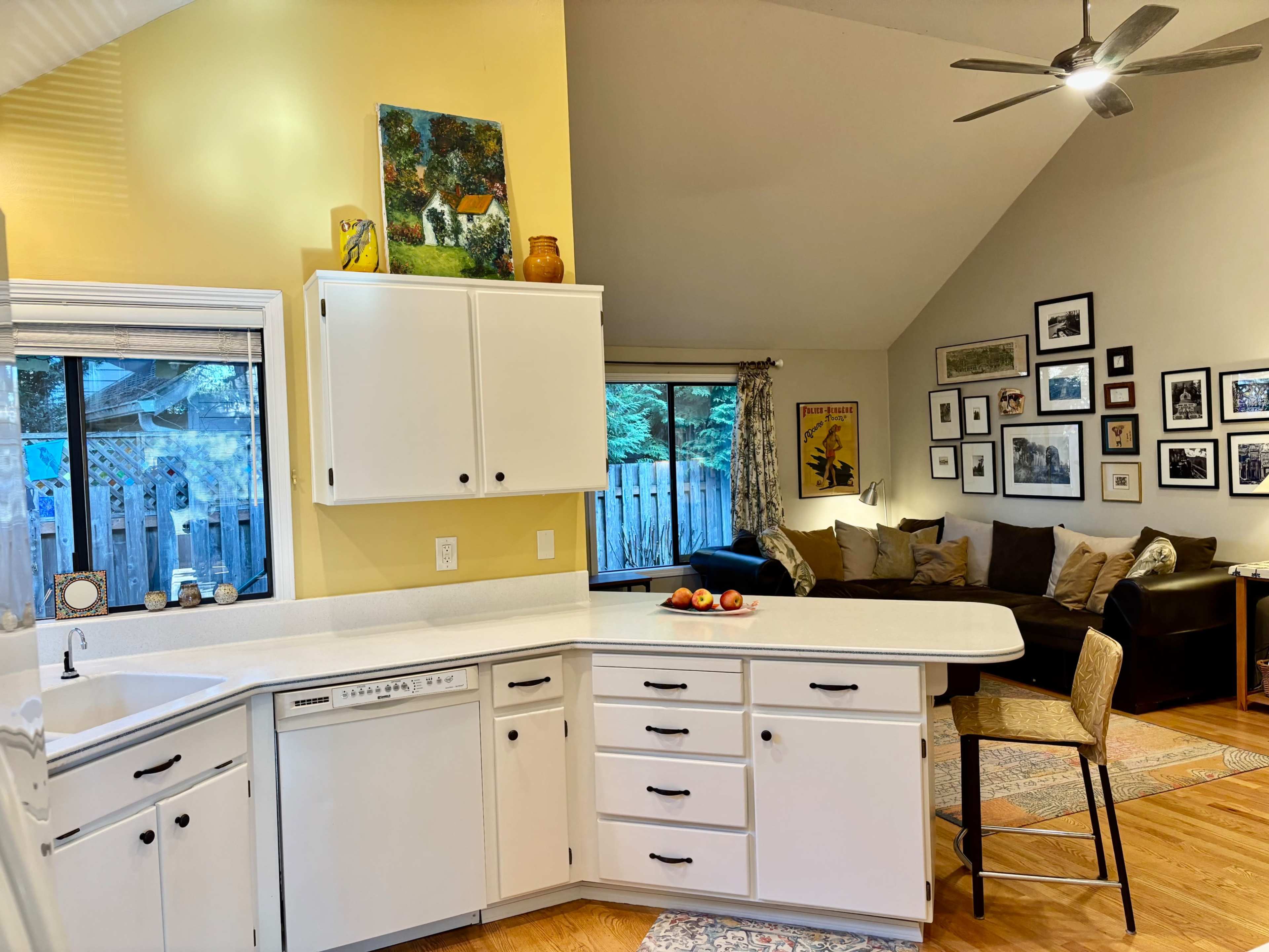The image shows a bright kitchen with a white countertop and cabinets that opens into a cozy living area featuring a sectional sofa and framed artwork on the walls.