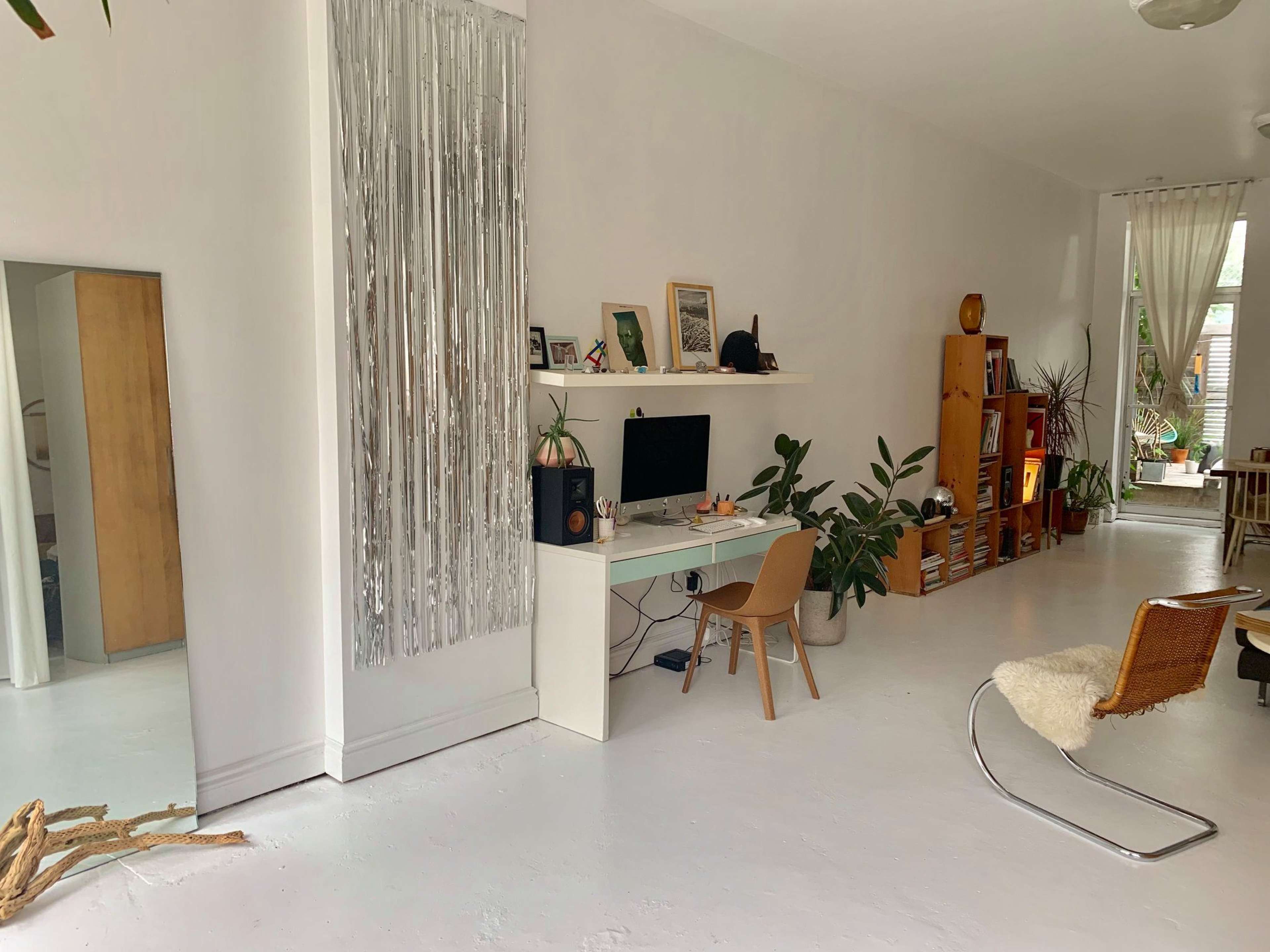 A bright, minimalistic living space features a workspace against a white wall, plants, wooden shelves filled with books, and a light-colored floor.