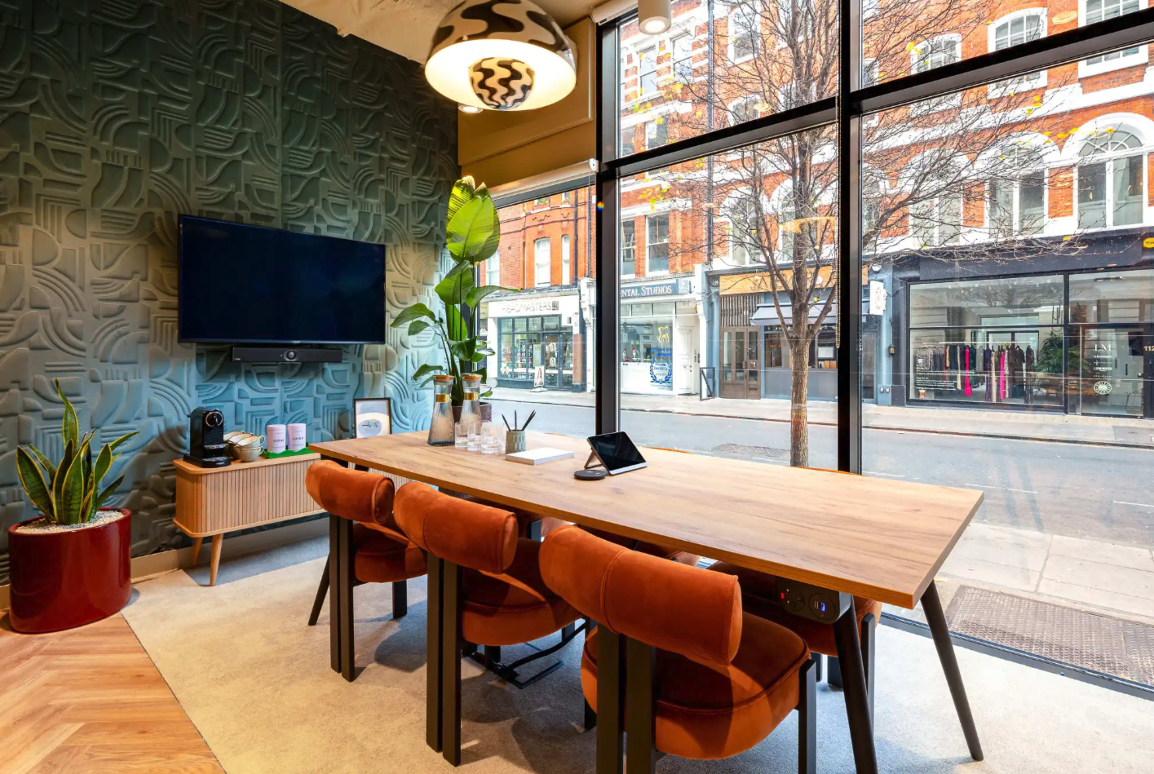 A modern conference room features a large wooden table surrounded by orange chairs, plants, and a wall-mounted television, with large windows overlooking a city street.