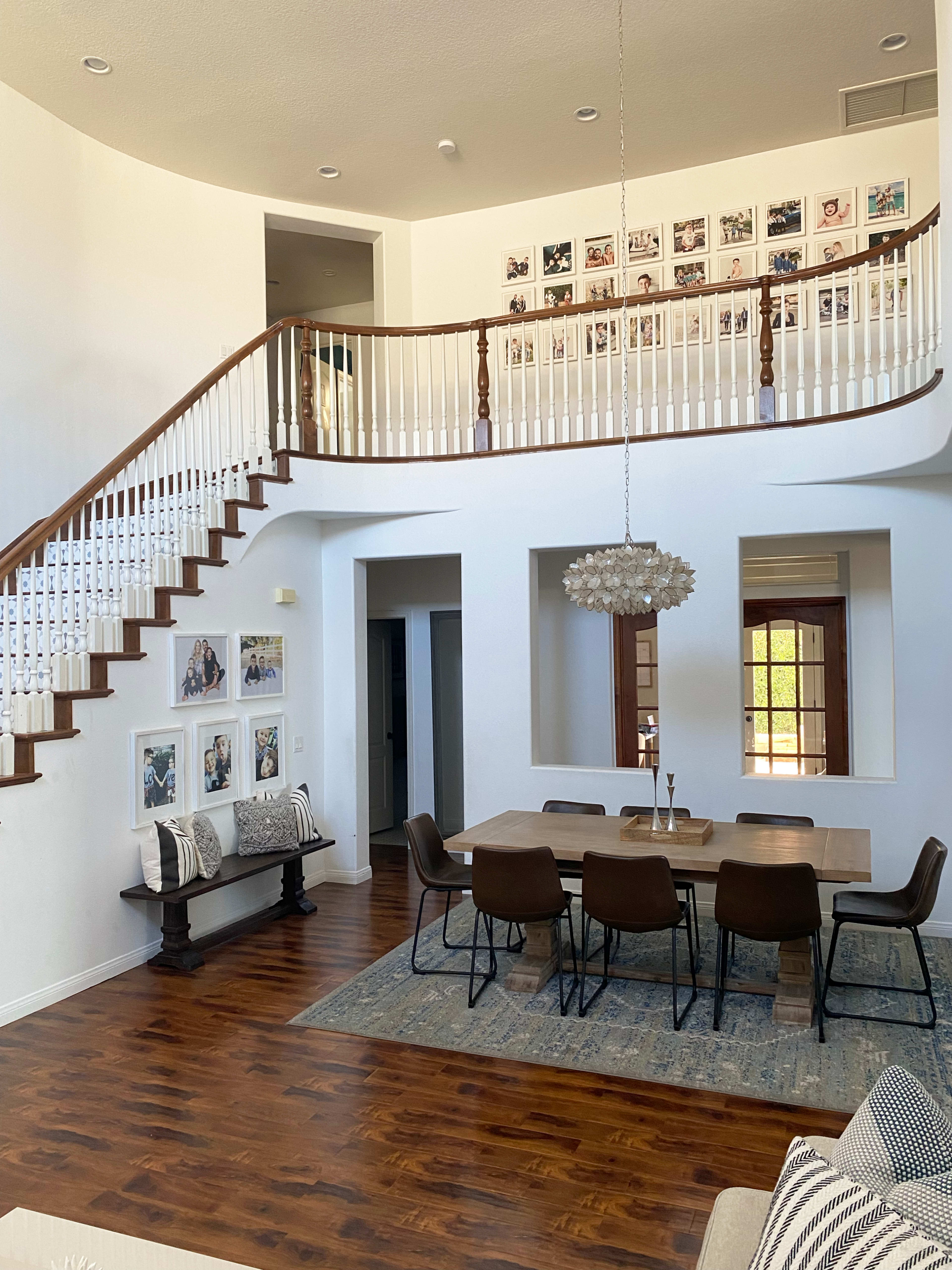 The image shows a modern, spacious dining area with a long wooden table, surrounded by chairs, and a staircase leading to an upper level adorned with framed photos.