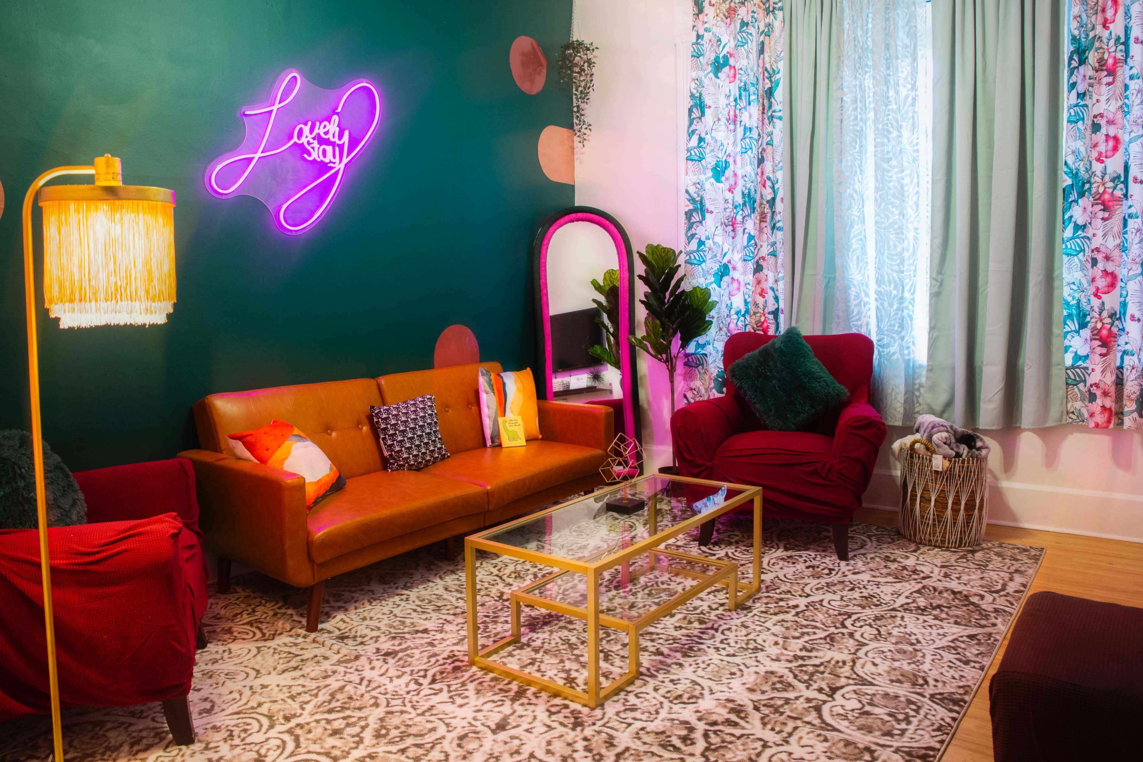 A cozy living room features a vibrant green wall, a neon sign, a brown leather sofa, and decorative curtains near a window.