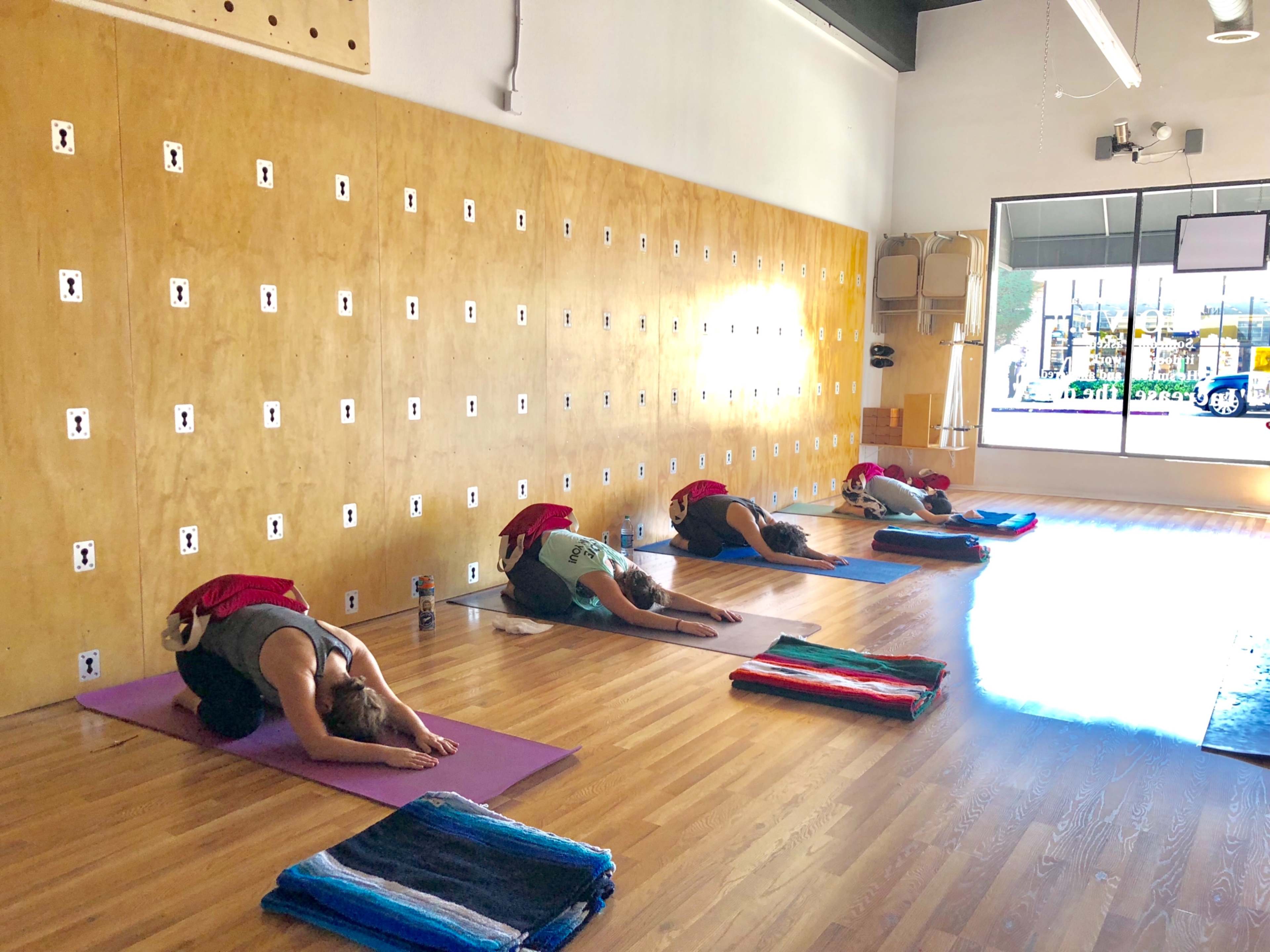 A yoga studio with several individuals practicing a pose on mats, positioned in a row along a wooden floor, against a wall with various hooks.