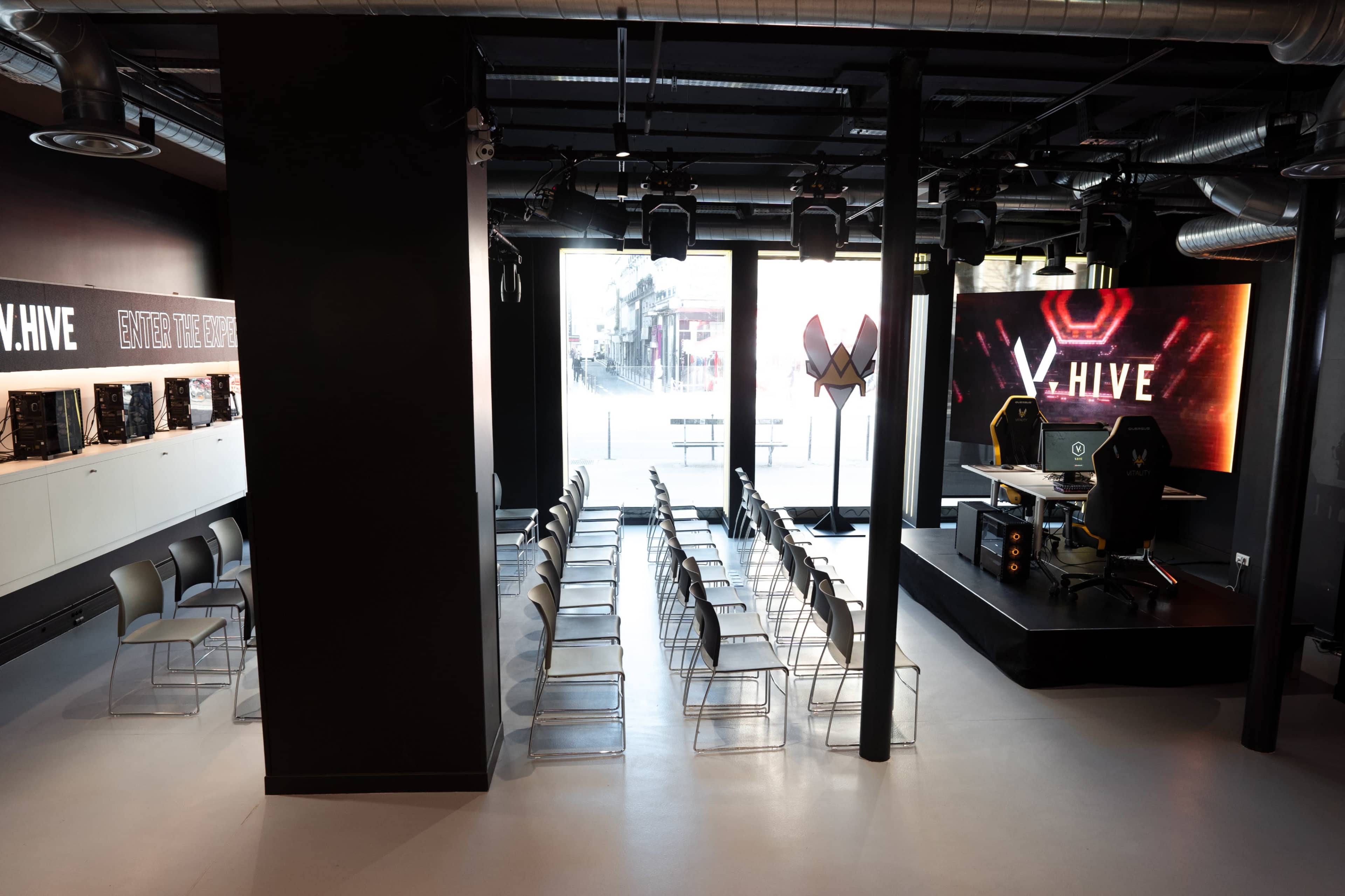 The image shows a modern indoor space set up for an event, featuring rows of clear chairs facing a stage with a large display screen and equipment.