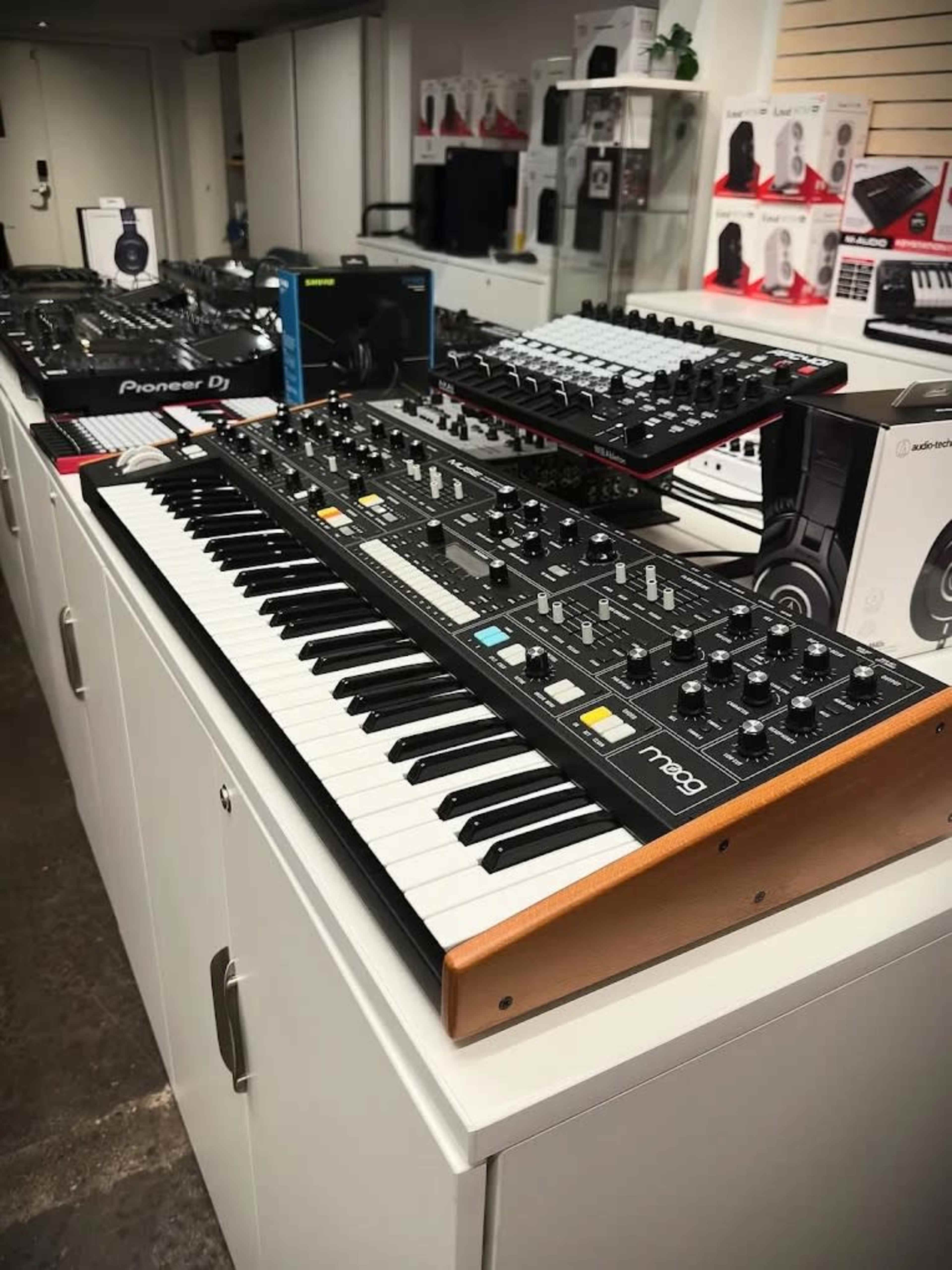 A row of music equipment, including a synthesizer, headphones, and audio interfaces, is displayed on a white countertop in a store.