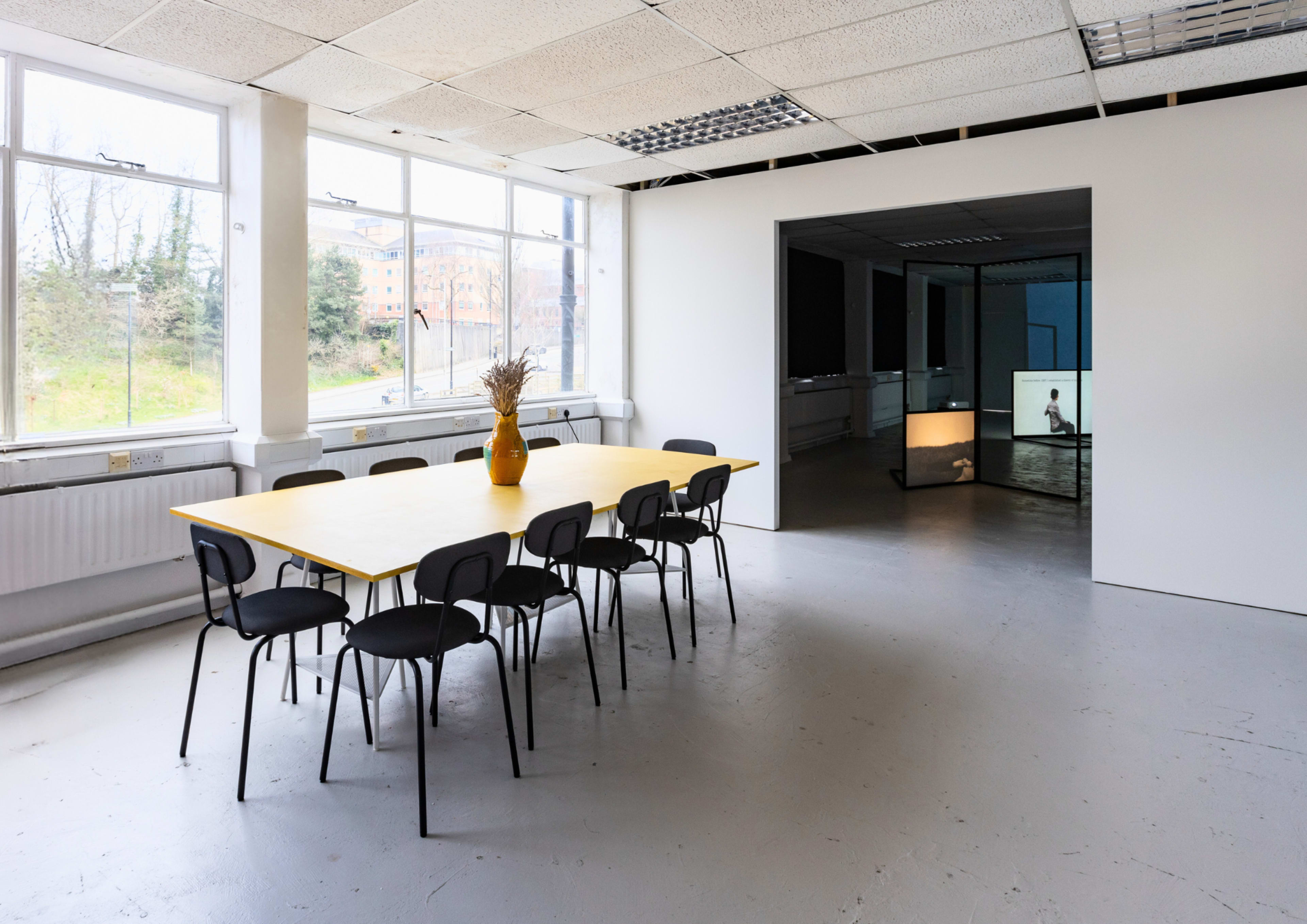 The image shows a spacious office interior with a large yellow table surrounded by black chairs and a view of a hallway leading to another room.
