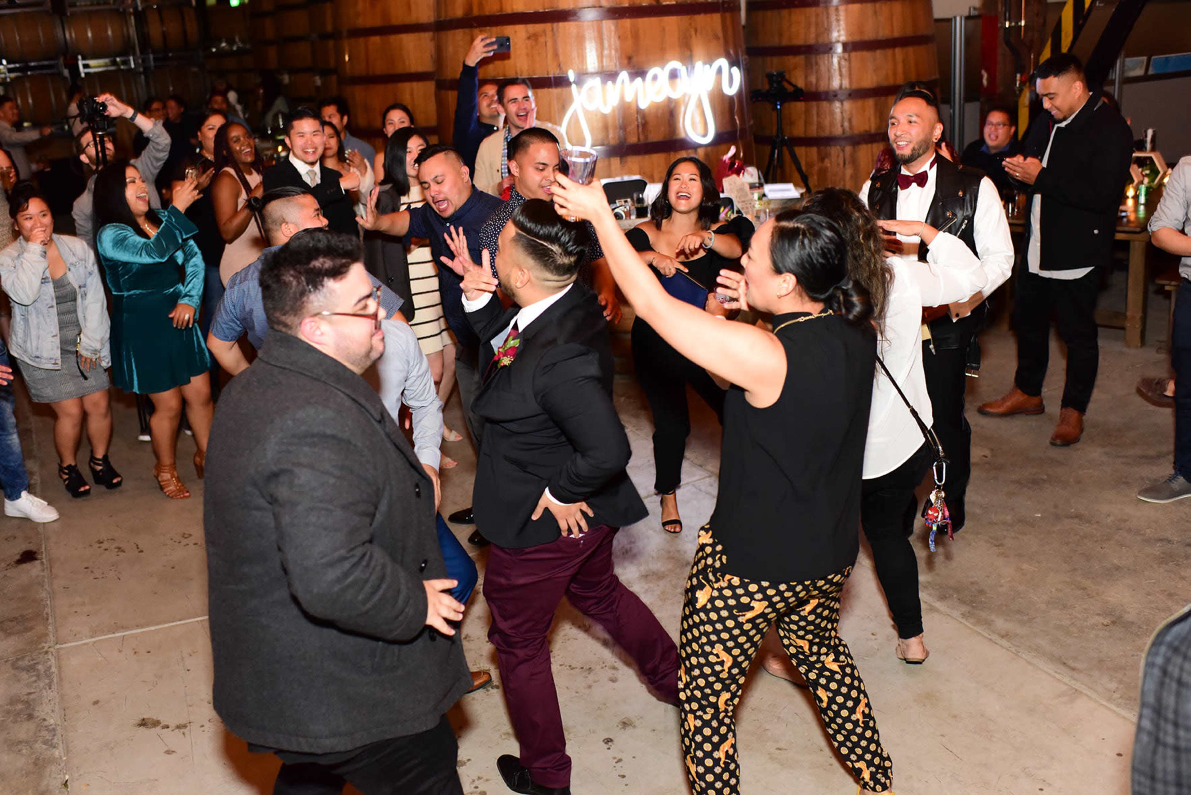 A lively group of people dances together in a large room with wooden barrels in the background.