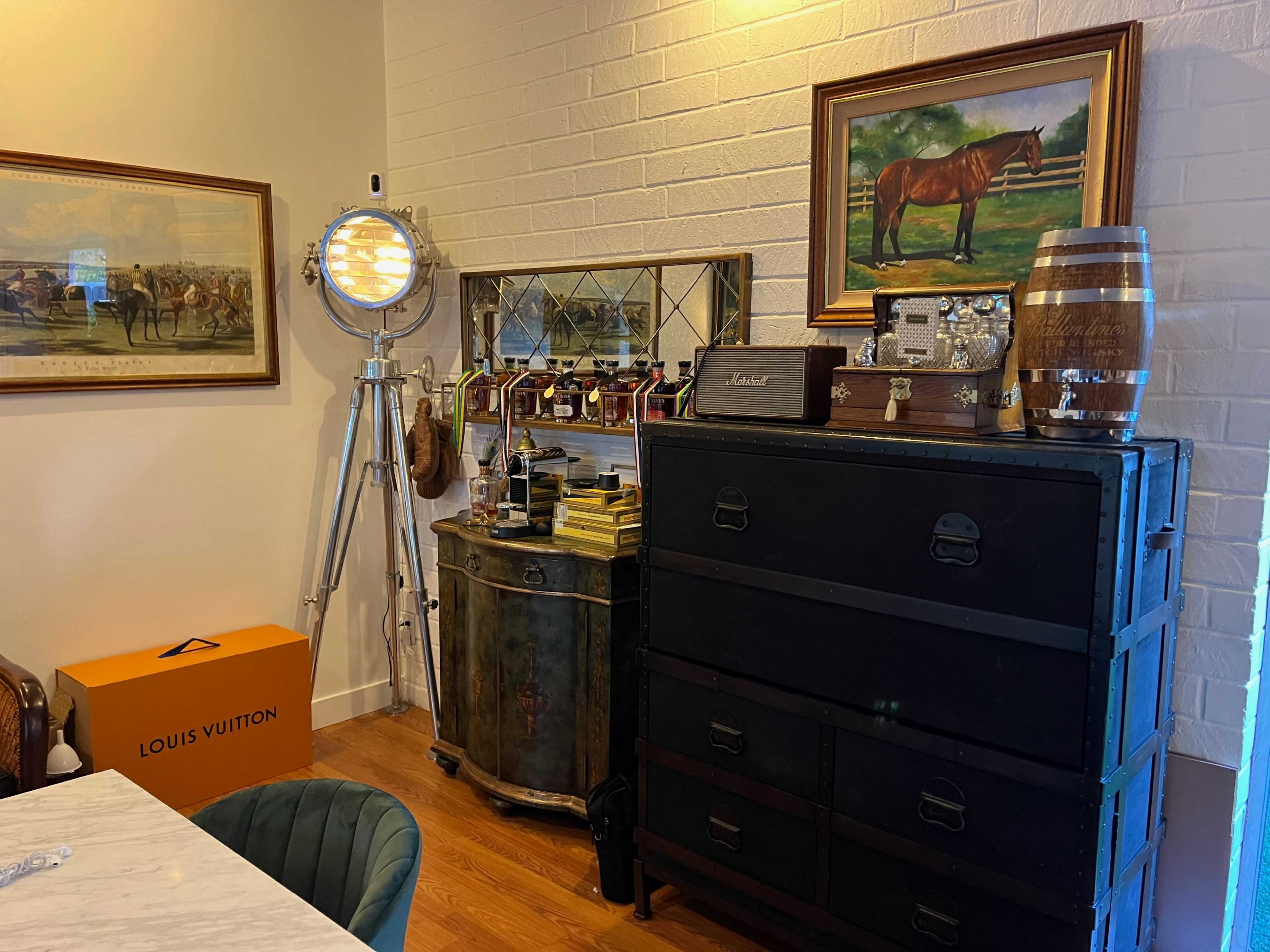 A room with vintage decor, including a horse painting, a metal cabinet, a Louis Vuitton box, and a film-style floor lamp.
