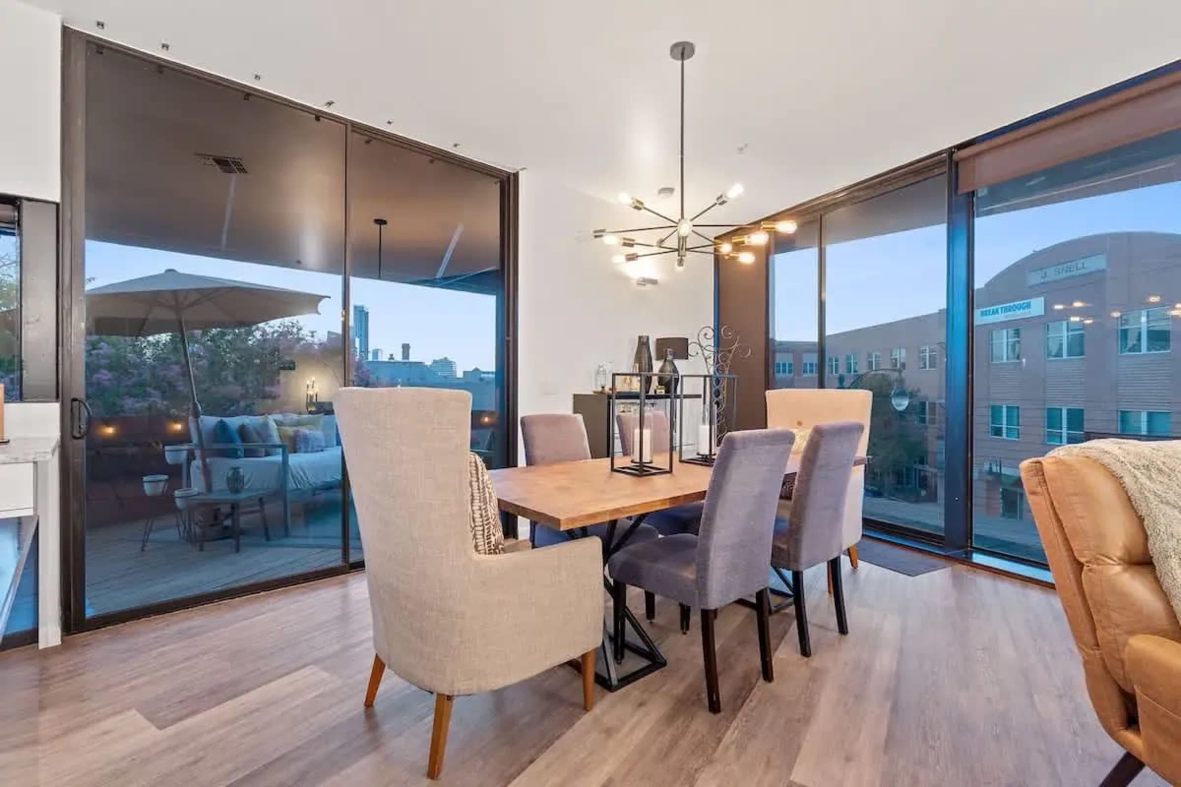 A dining area features a wooden table surrounded by six upholstered chairs, with large glass windows showcasing a balcony view.