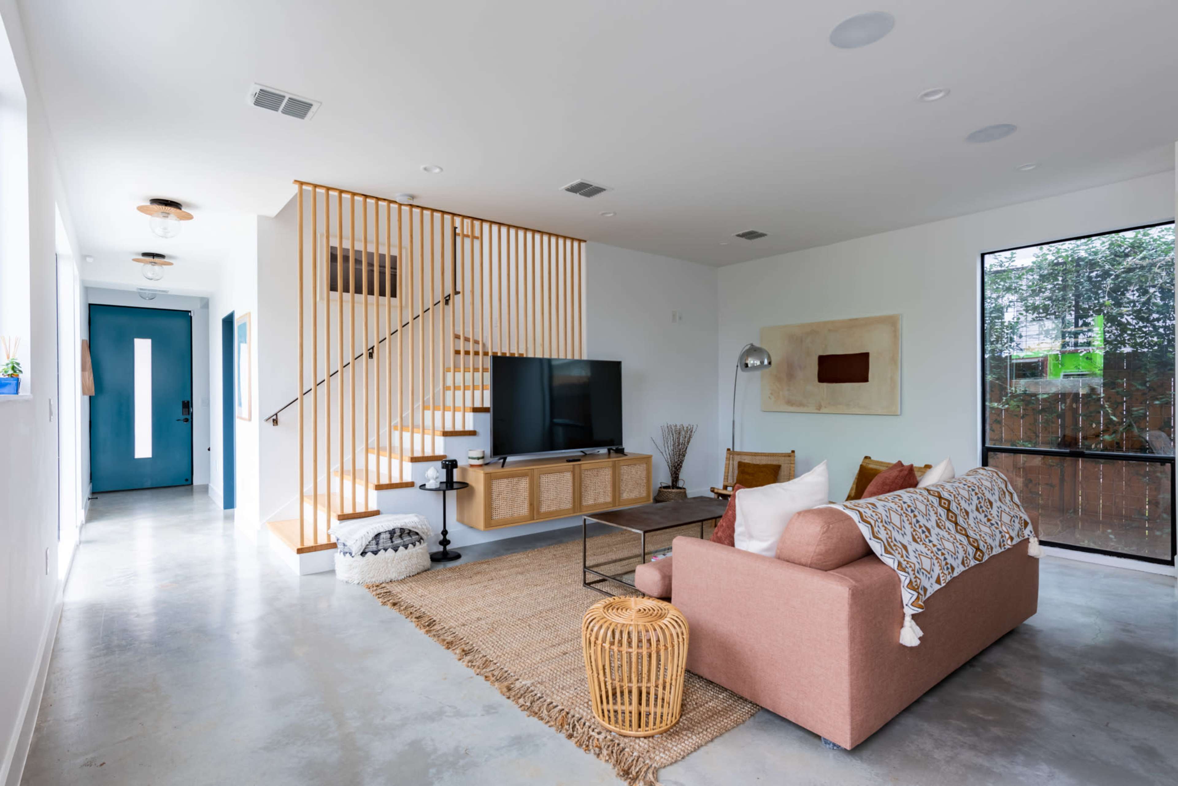The image shows a spacious living room with a pink sofa, a wooden TV stand, and a staircase leading to an upper level, complemented by a hallway and large windows.