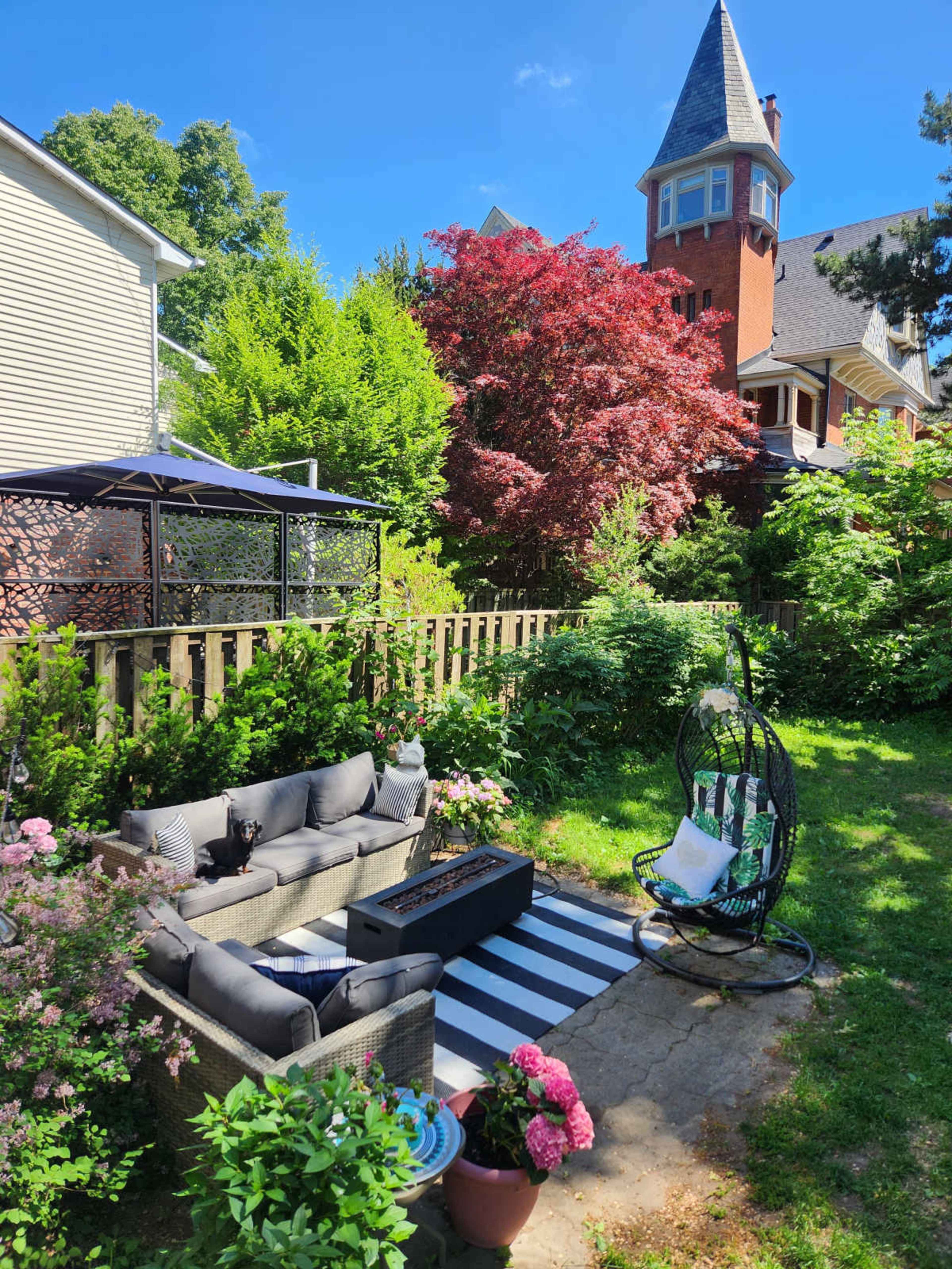 The image shows a landscaped backyard with a seating area featuring a sectional couch, a striped rug, and a hanging chair, surrounded by greenery and flowering plants.