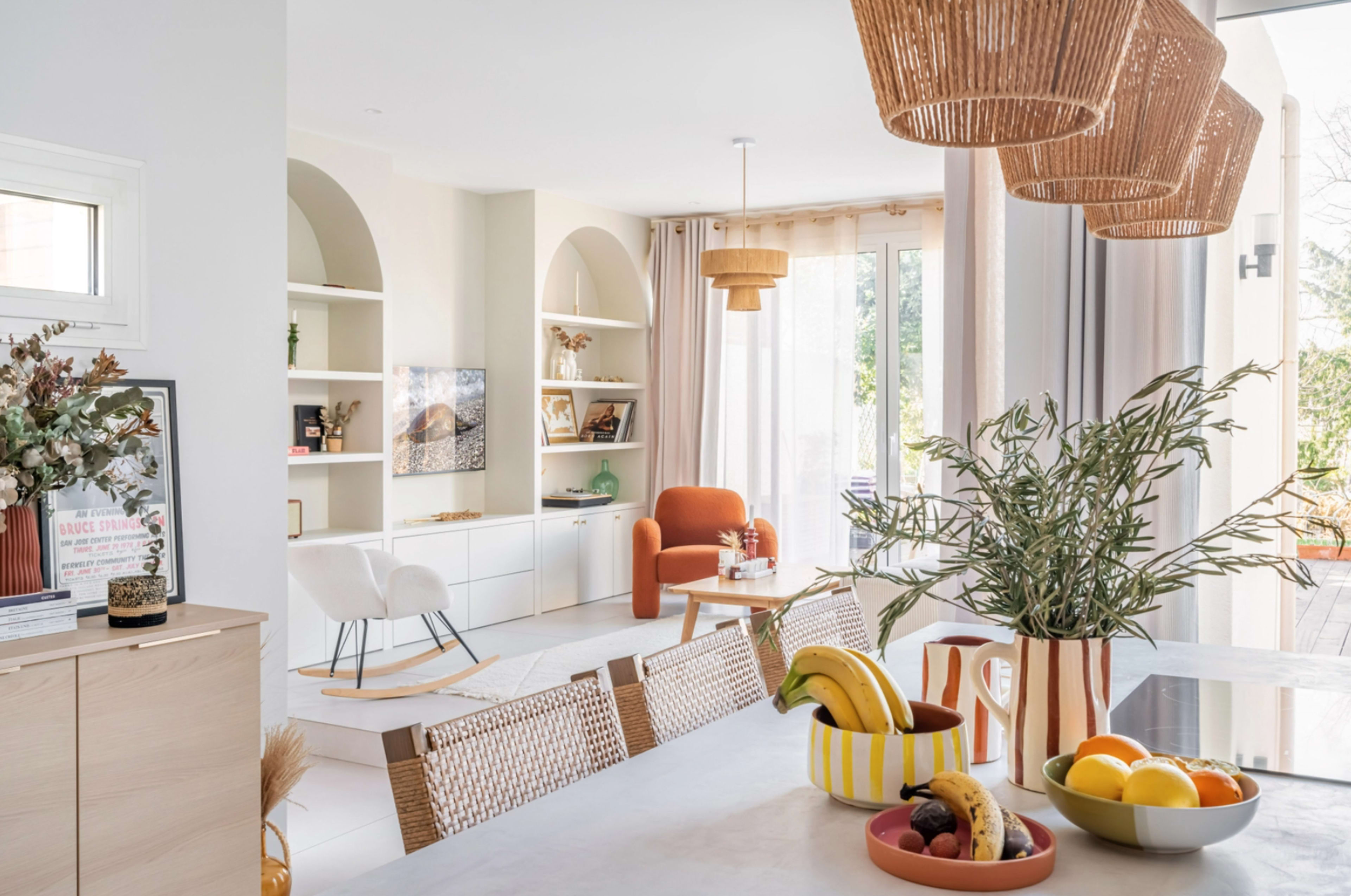 A bright, modern living space with open shelving, a dining area featuring colorful bowls of fruit, and stylish pendant lights overhead.