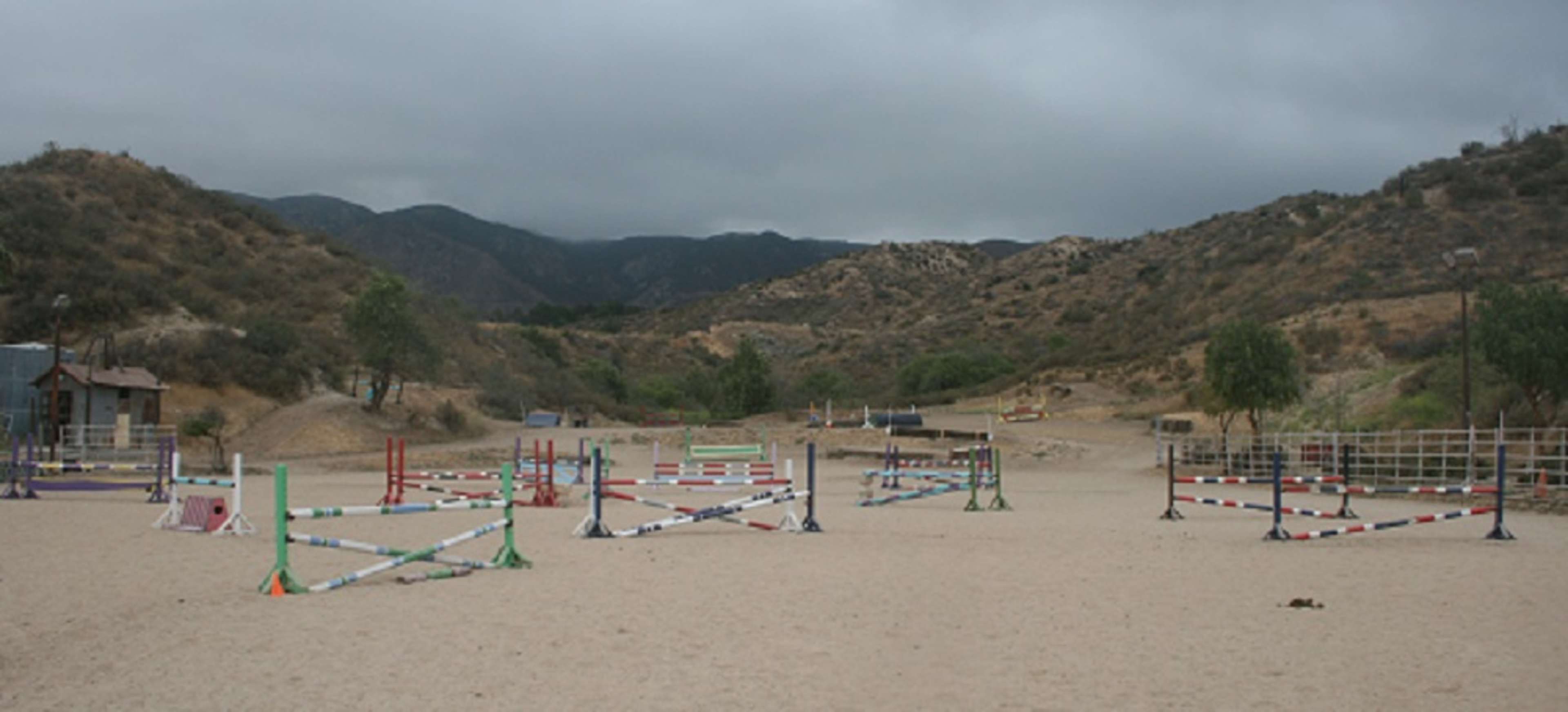 The image shows an equestrian arena with multiple colored jumps set up on sandy terrain surrounded by hills and overcast skies.