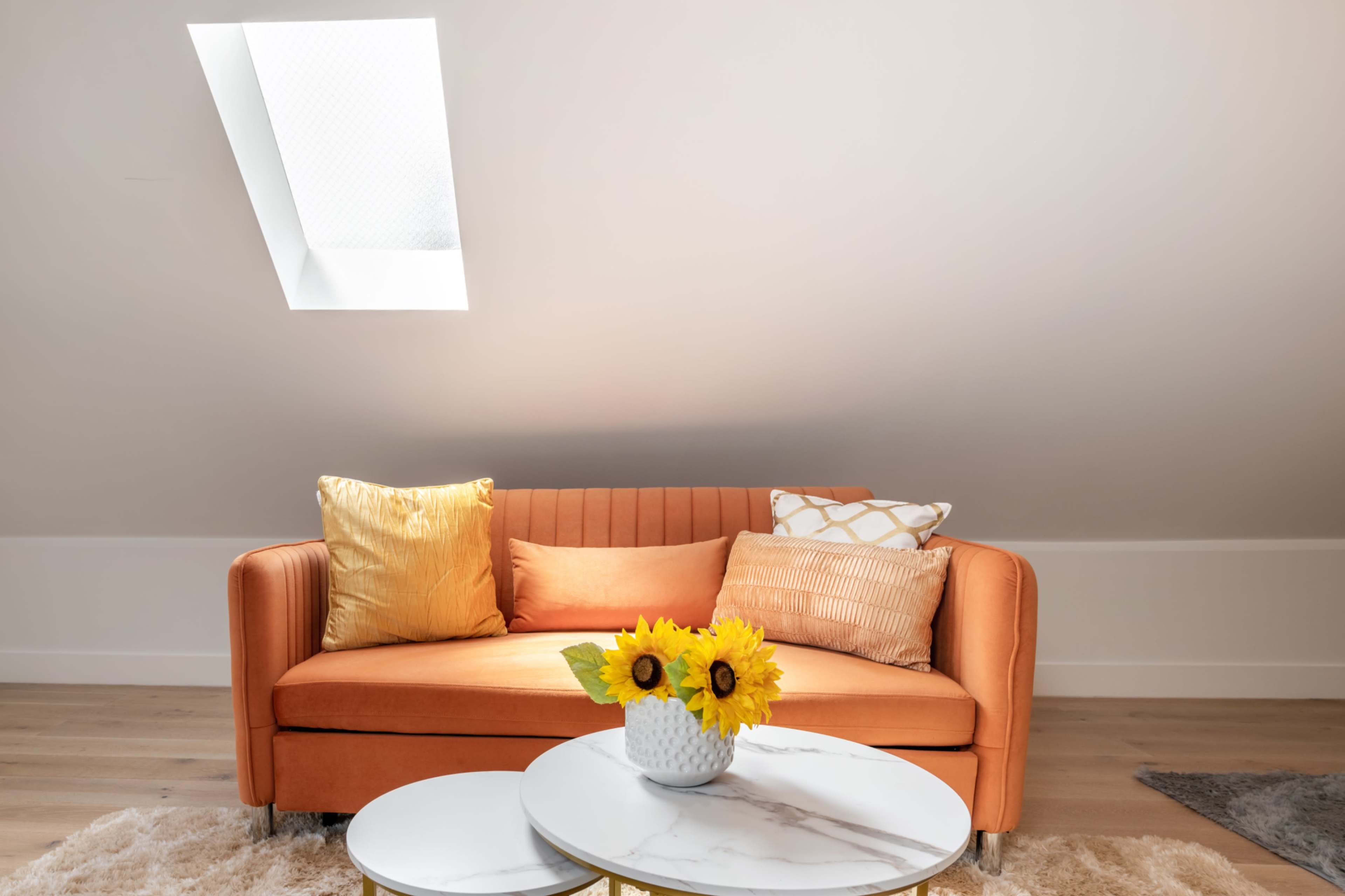 A bright room features an orange sofa adorned with cushions and a small white table holding a vase of sunflowers.