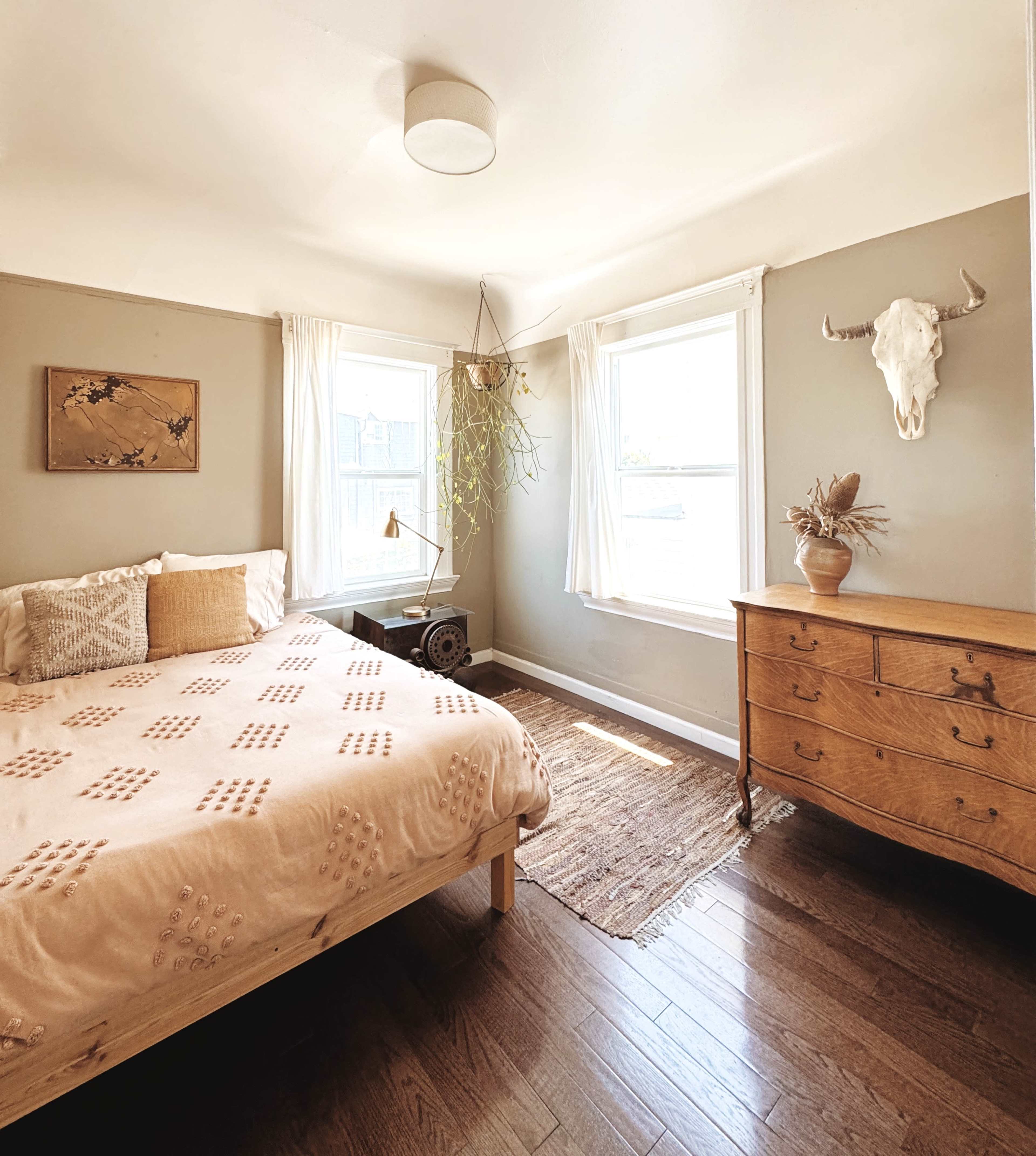 The image shows a bright bedroom featuring a bed with a pink patterned duvet, a wooden dresser, two windows with sheer curtains, and a decorative wall hanging.