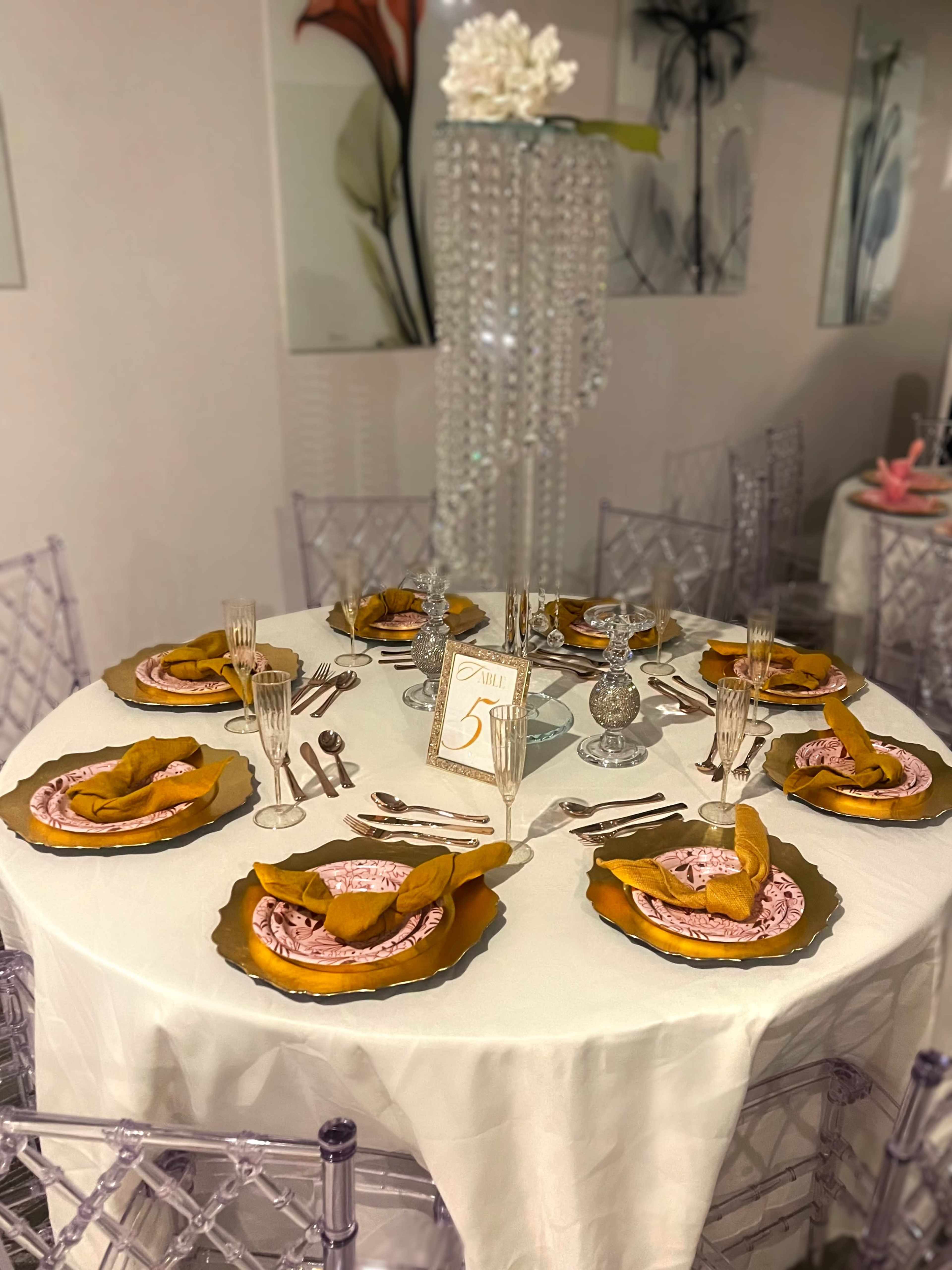A round dining table is set with elegant plates, silverware, and woven napkins, decorated with a flower centerpiece and a small table number.