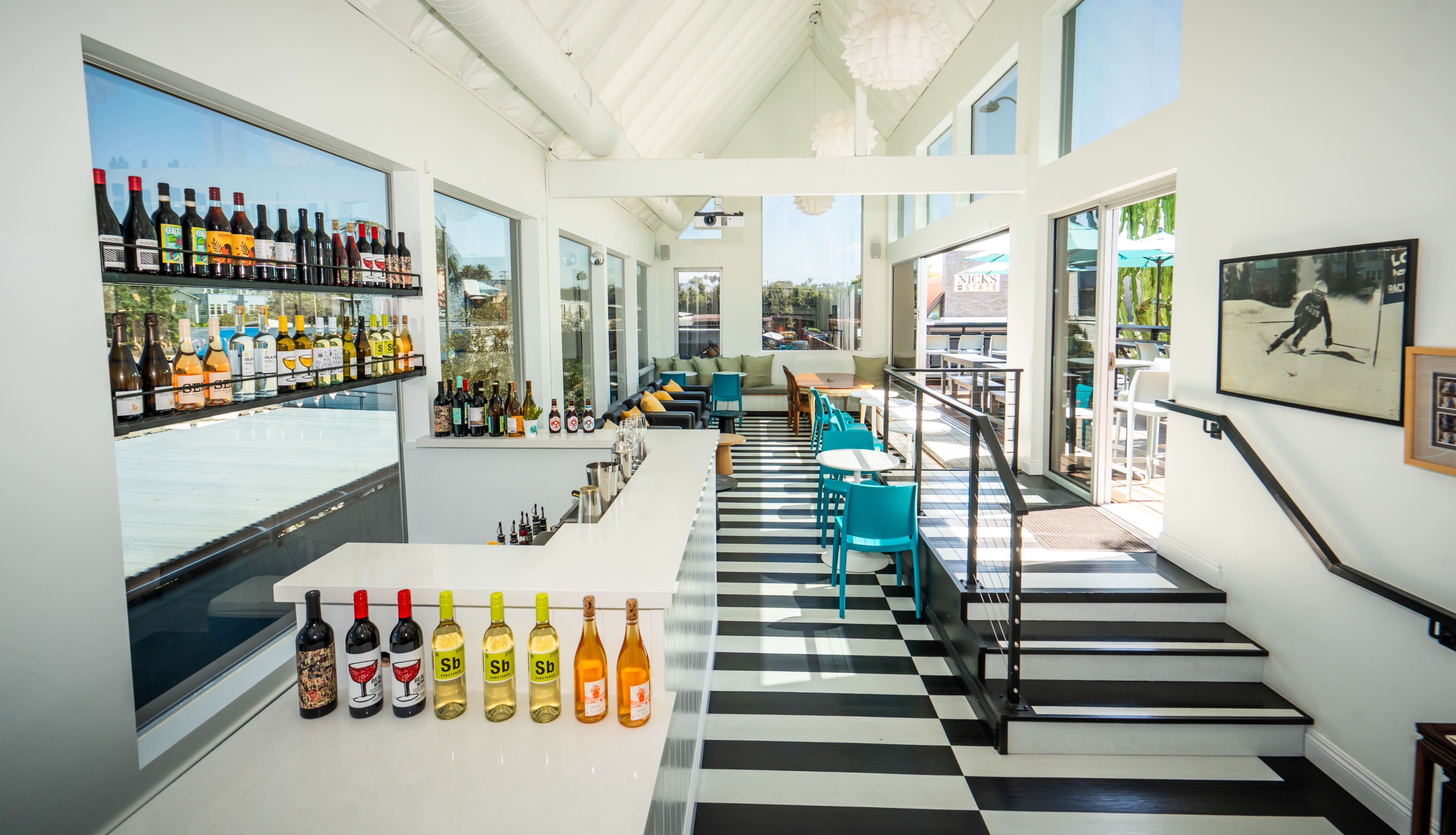 The image shows a modern bar area with a white countertop, a variety of colorful liquor bottles displayed on shelves, and an open seating space with blue chairs and natural light coming through large windows.