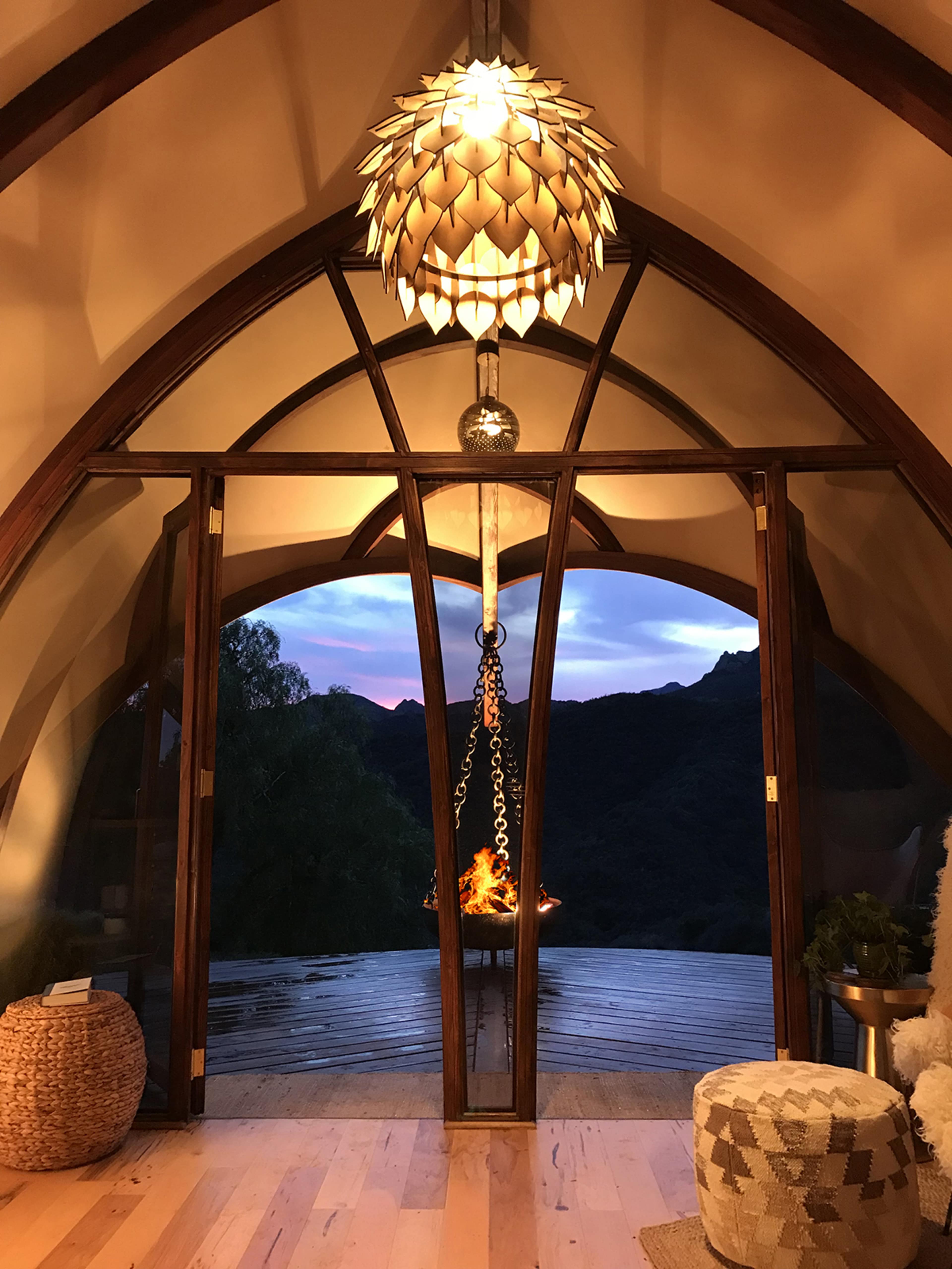 The image shows a cozy interior with a unique pendant light and large open doors leading to a wooden deck overlooking a mountainous landscape at sunset.