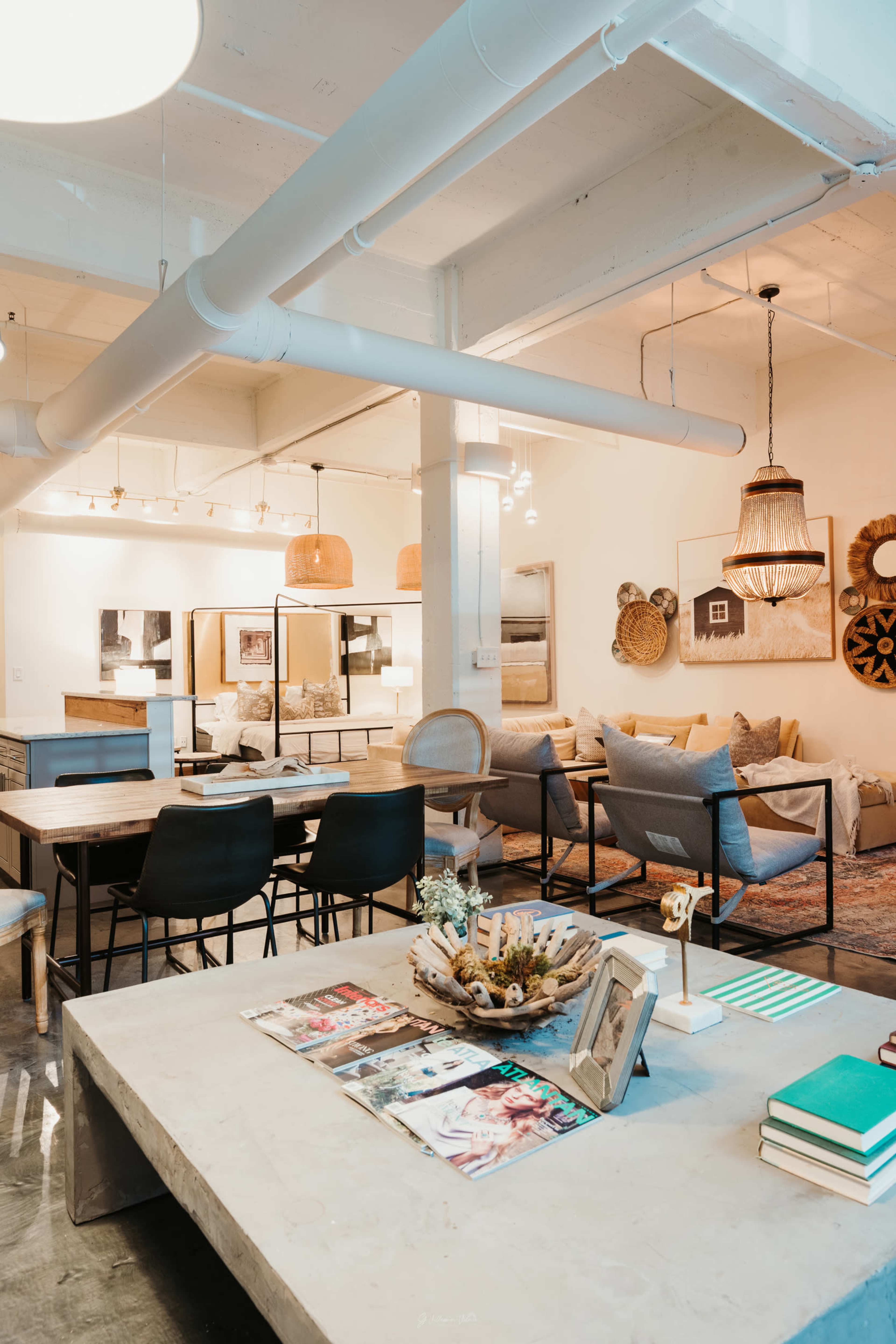 The image shows a modern open-concept space featuring a dining area with a large concrete table and black chairs, a lounge area with light gray sofas, and decorative items on display throughout.