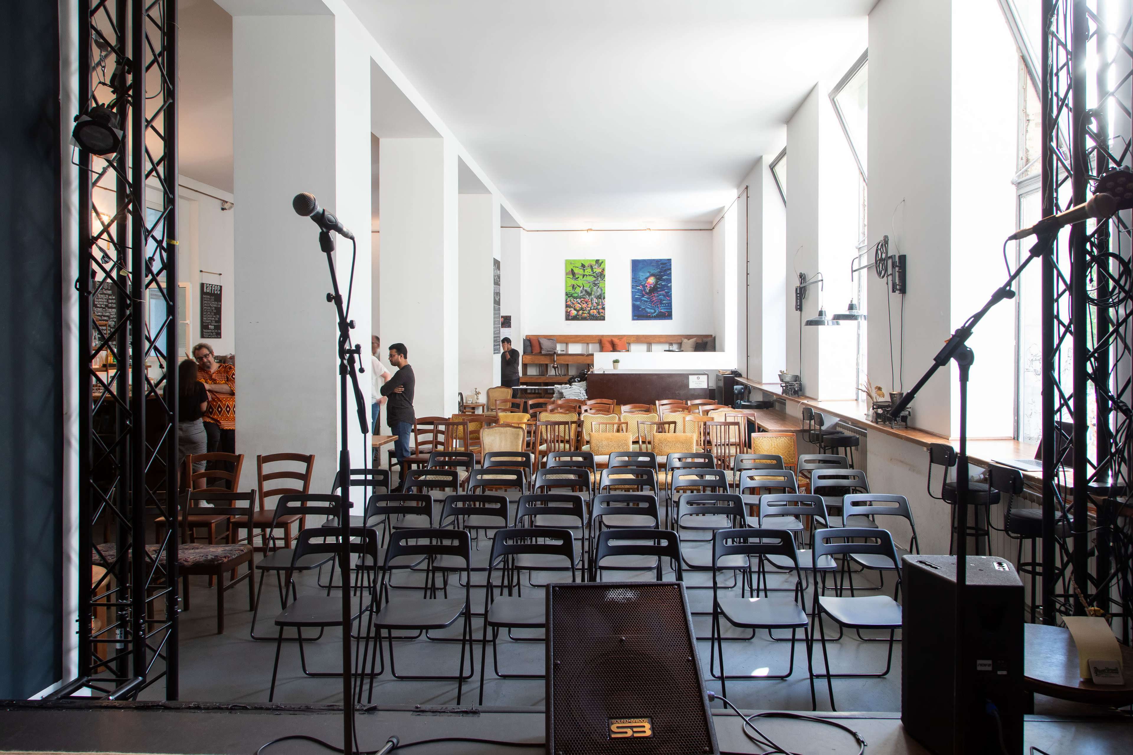 A sparsely furnished performance space with rows of black chairs facing a stage area, flanked by microphones and a bar in the background.