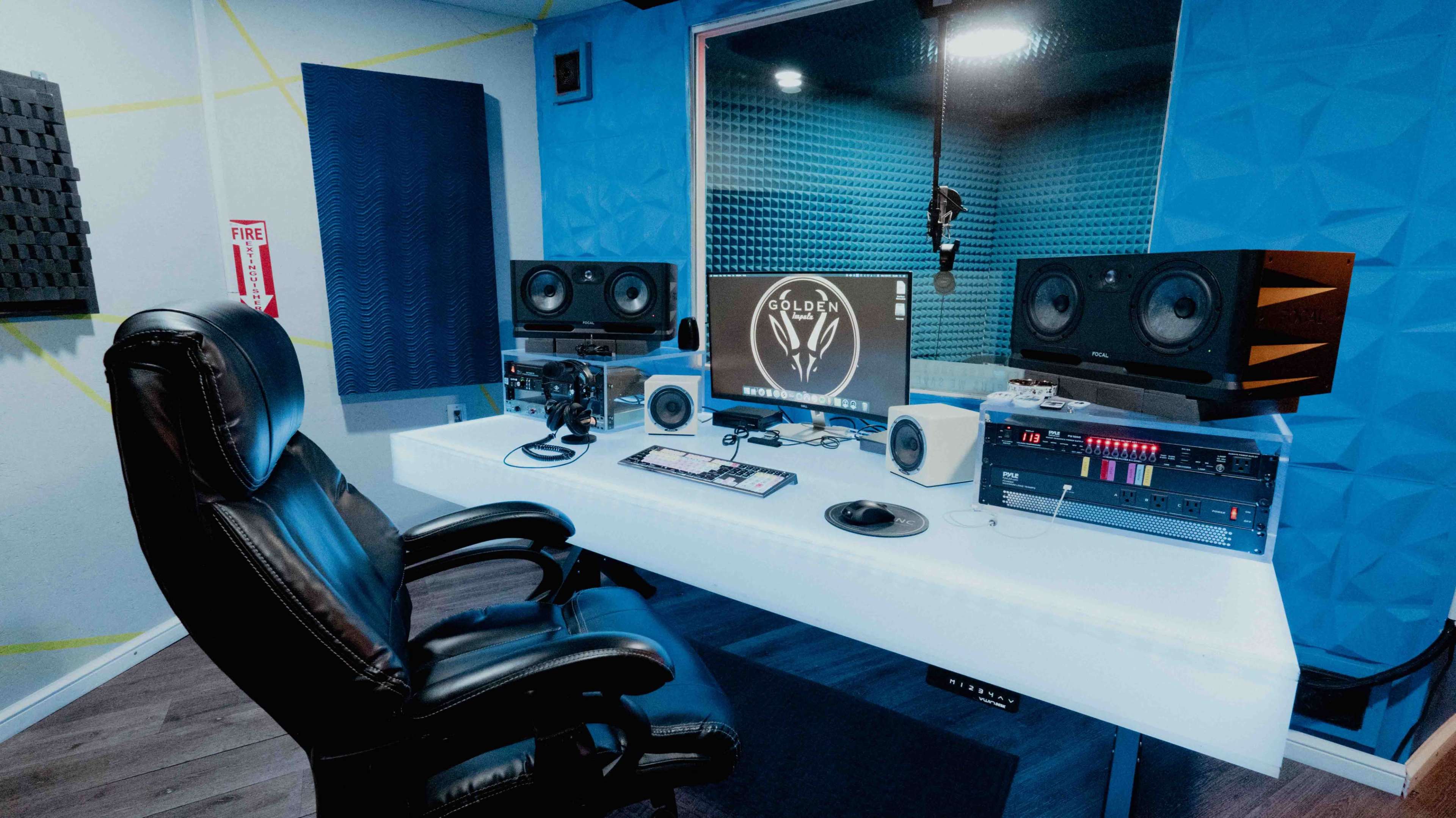The image shows a modern recording studio with a central desk containing audio equipment, speakers, a computer monitor, and an ergonomic chair.