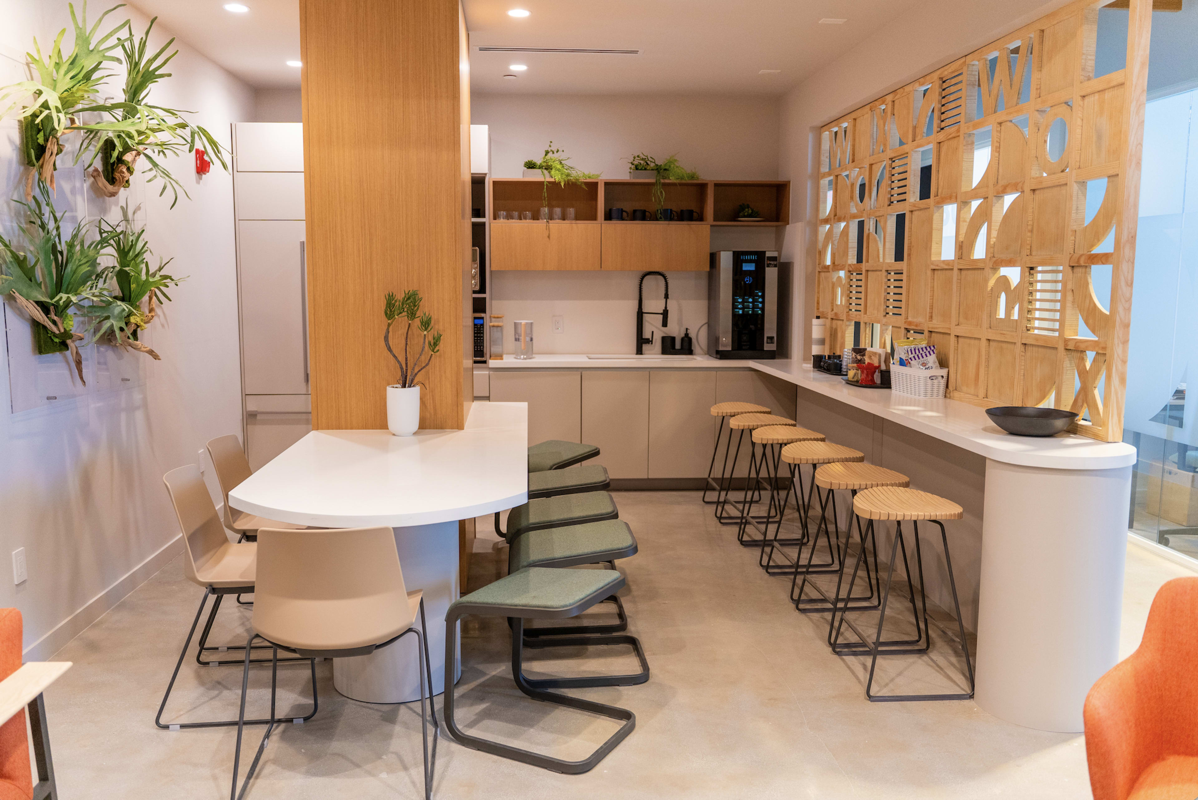 A modern kitchen area featuring a countertop with barstools, a dining table, and shelves with plants and appliances.