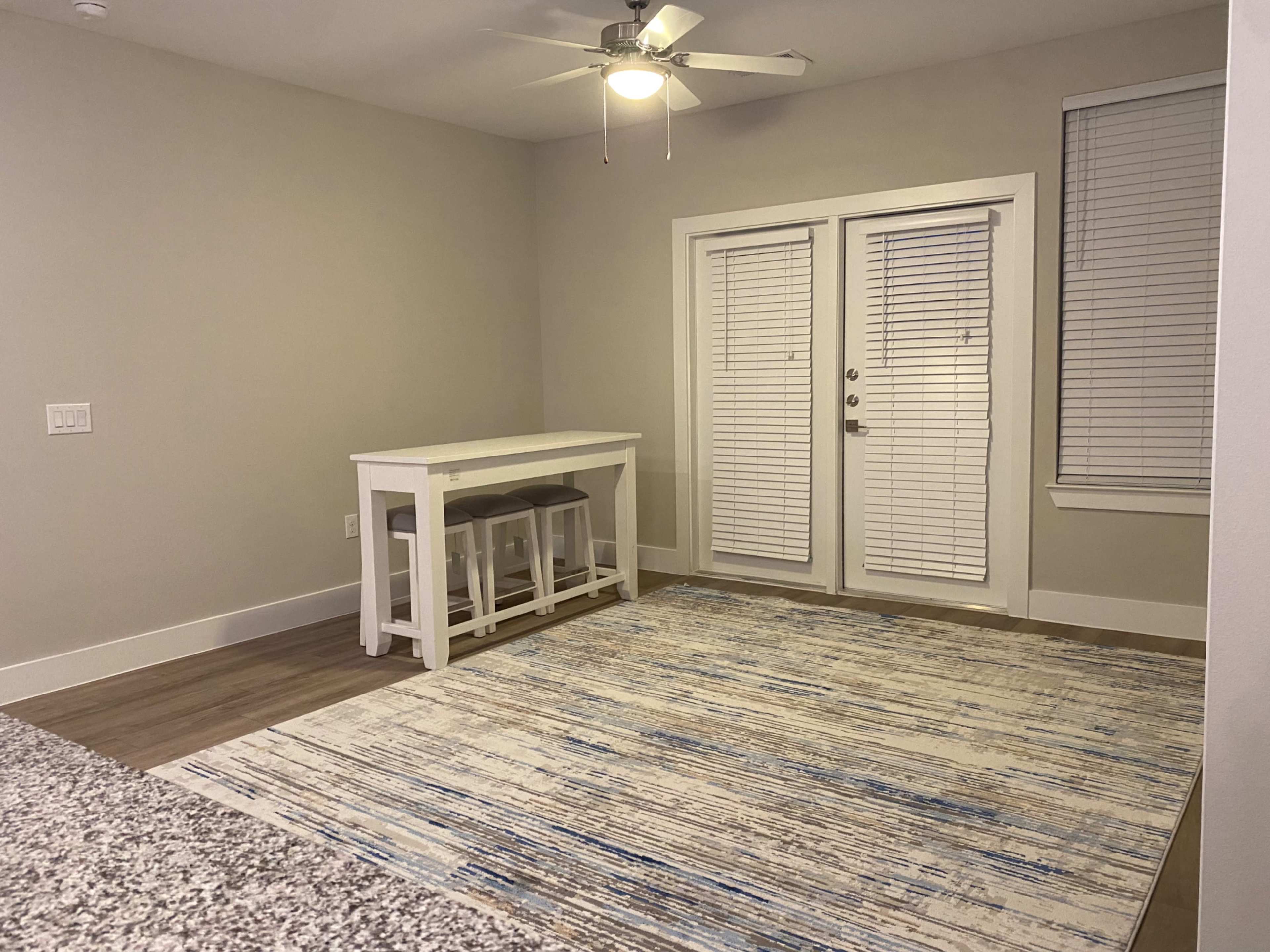 A spacious room featuring a small white dining table with three stools, a large area rug, and double glass doors leading to an outdoor space.