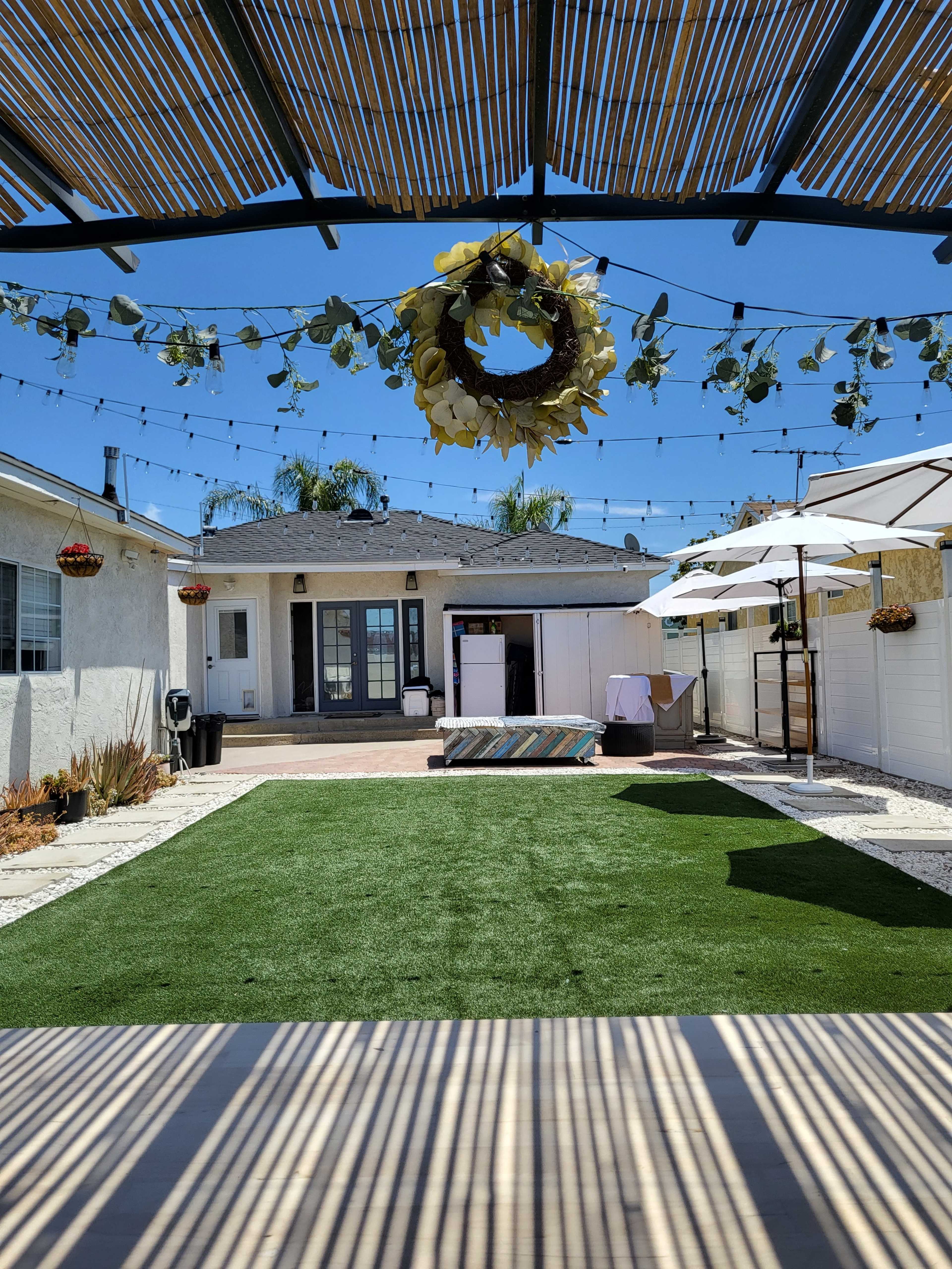 A backyard scene features a green lawn, a shaded area with string lights overhead, and a covered seating arrangement against the backdrop of a white house.
