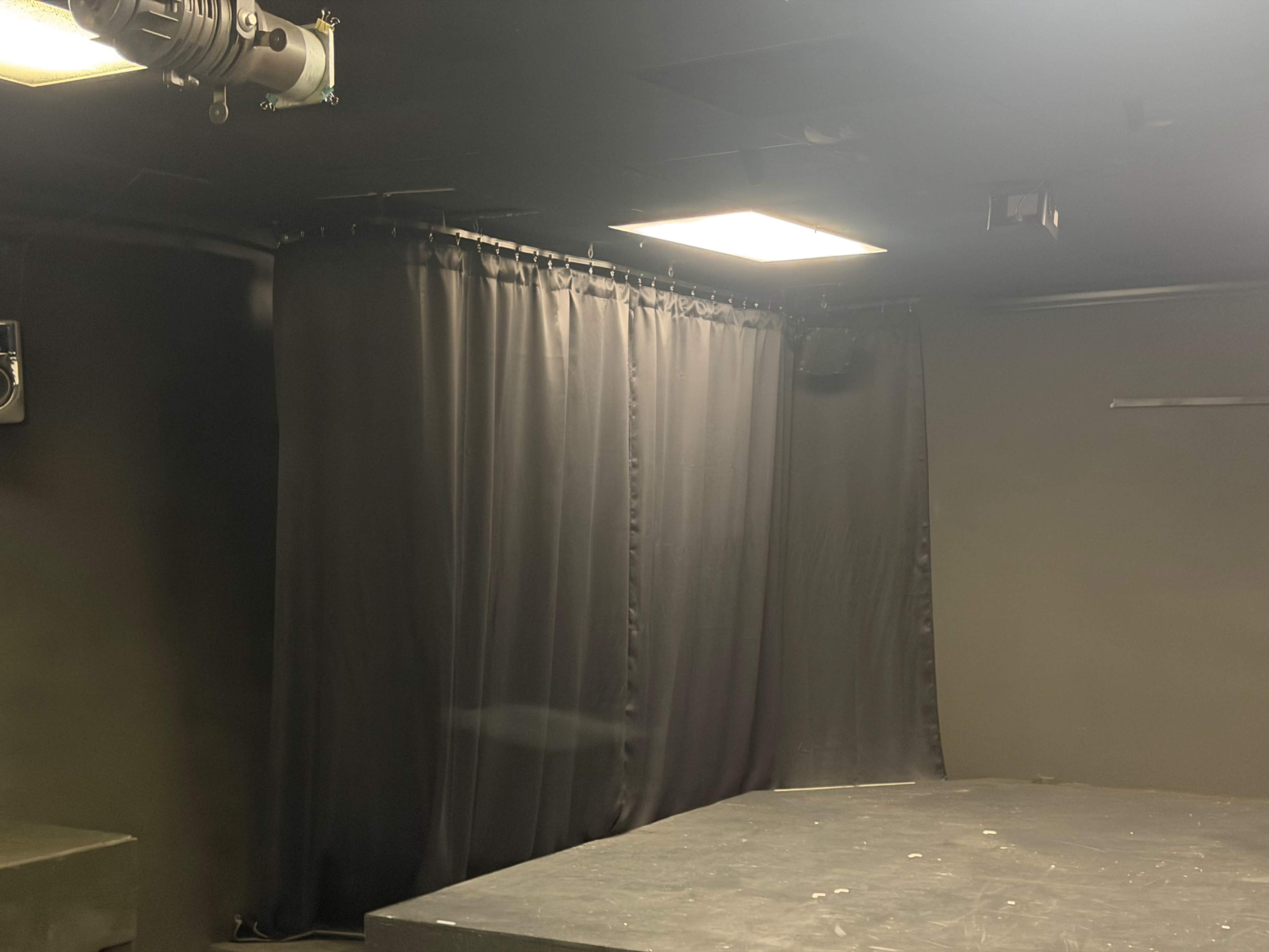 The image shows a dimly lit stage area with a black curtain and overhead lights in a theater setting.
