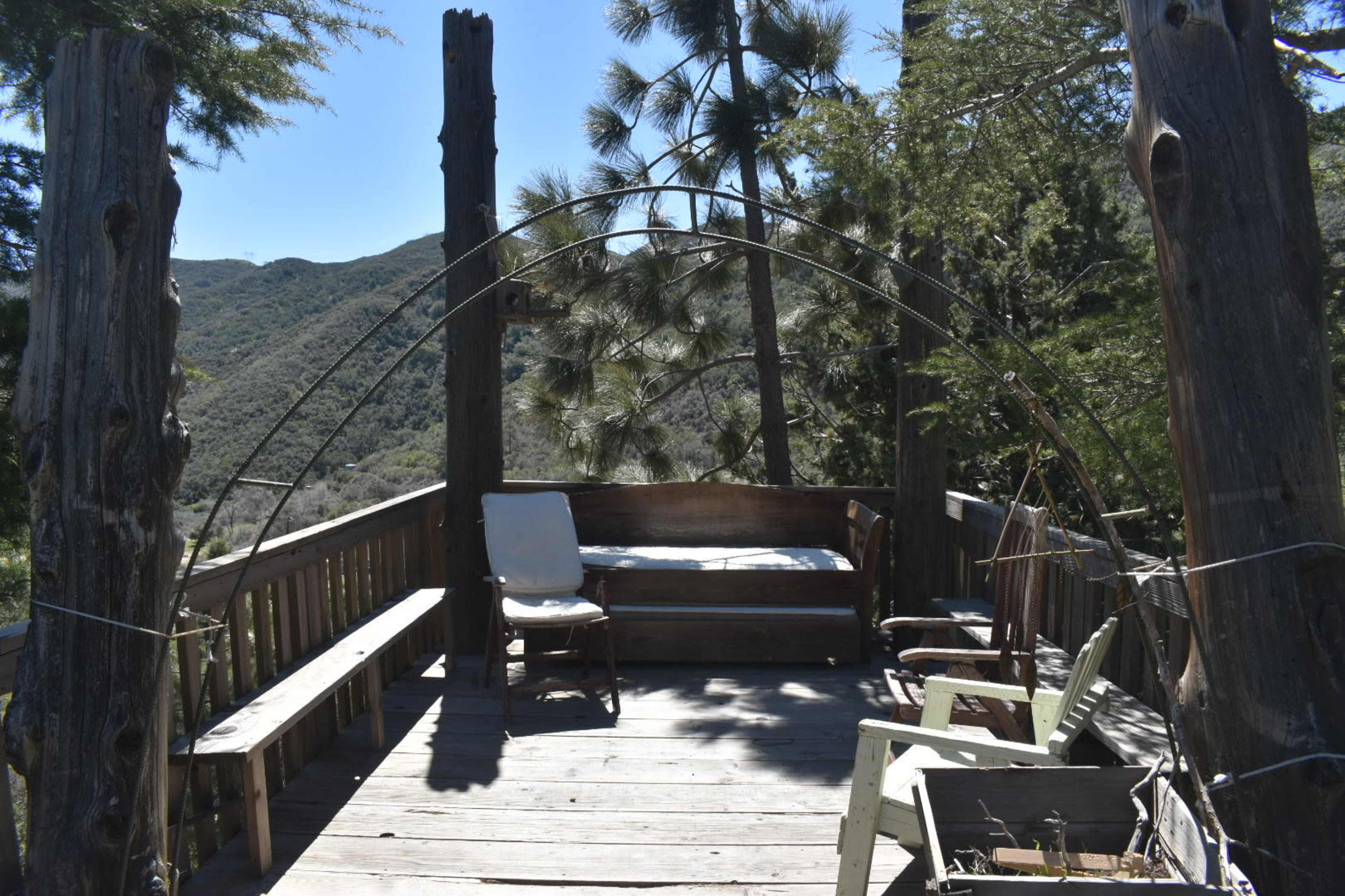 The image shows a wooden deck with rustic furniture surrounded by trees and hills in the background.
