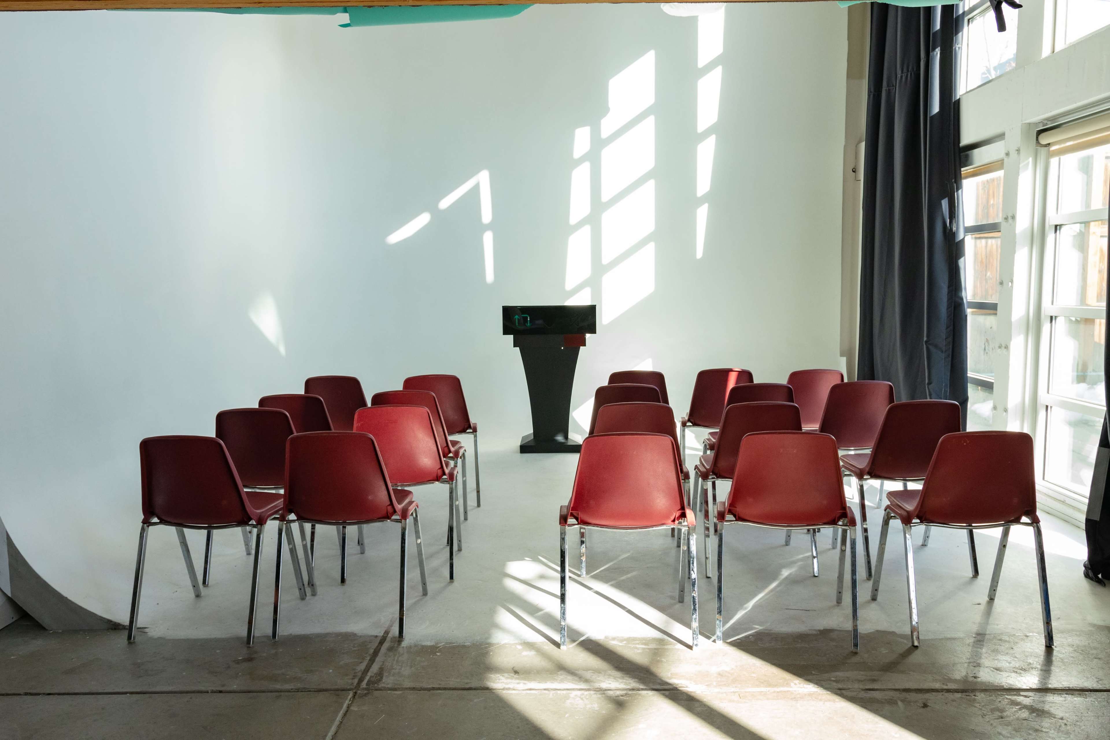 A row of red chairs is arranged in a semicircle around a podium in a bright, empty room with large windows.