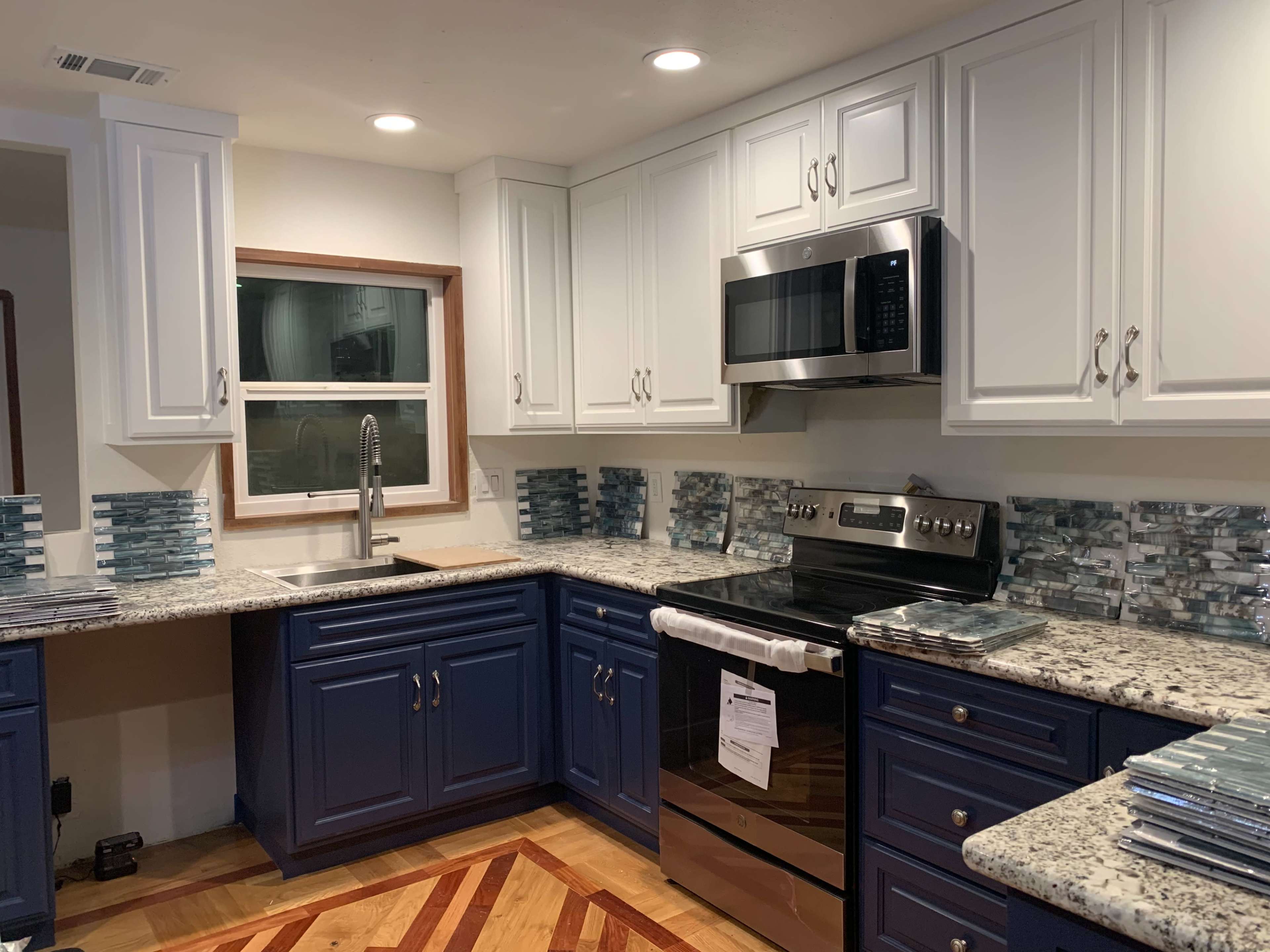 The image shows a modern kitchen with white upper cabinets, navy blue lower cabinets, stainless steel appliances, and a granite countertop with a patterned backsplash.