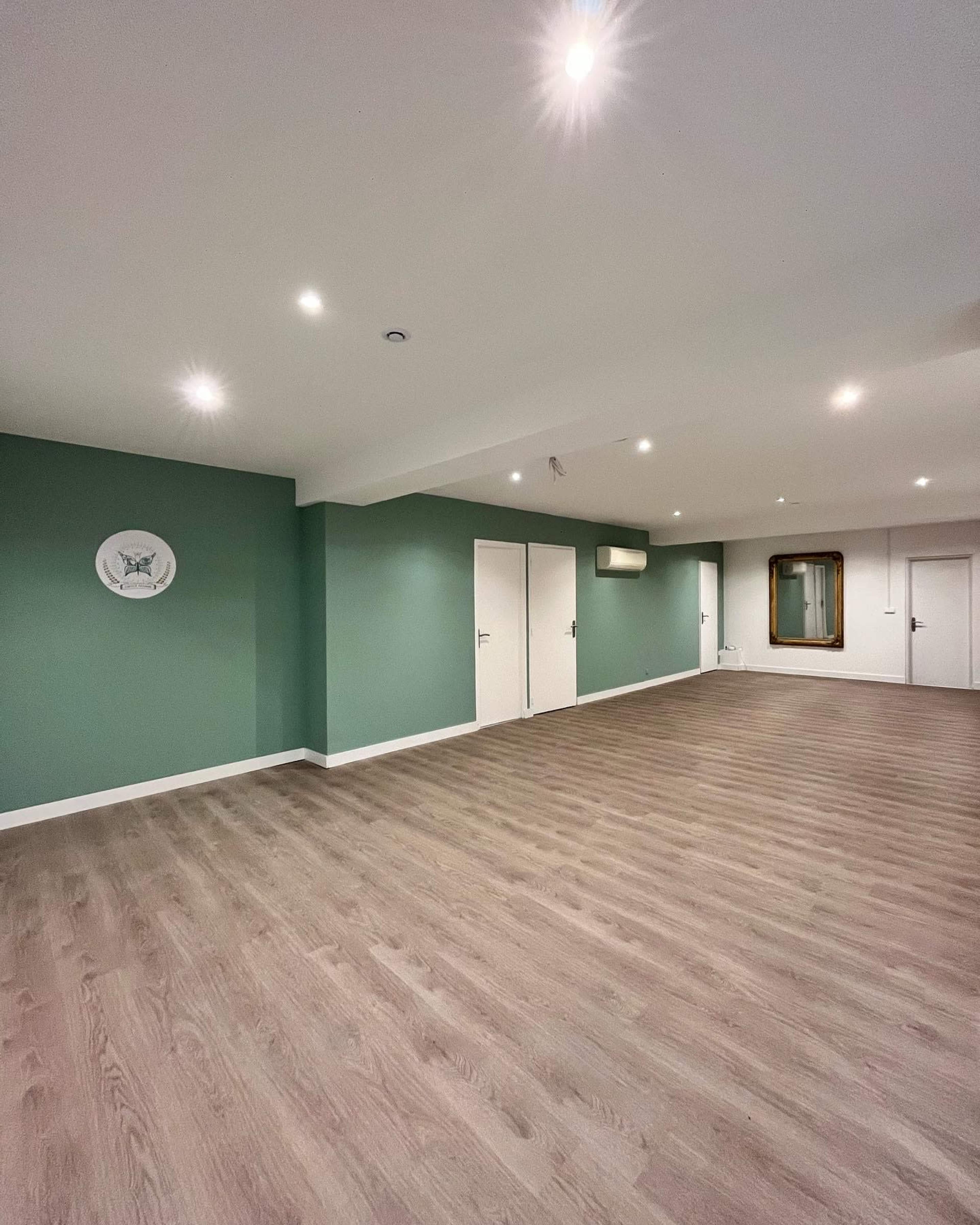 The image shows a spacious, empty room with green accent walls, hardwood flooring, recessed lighting, and two closed doors.