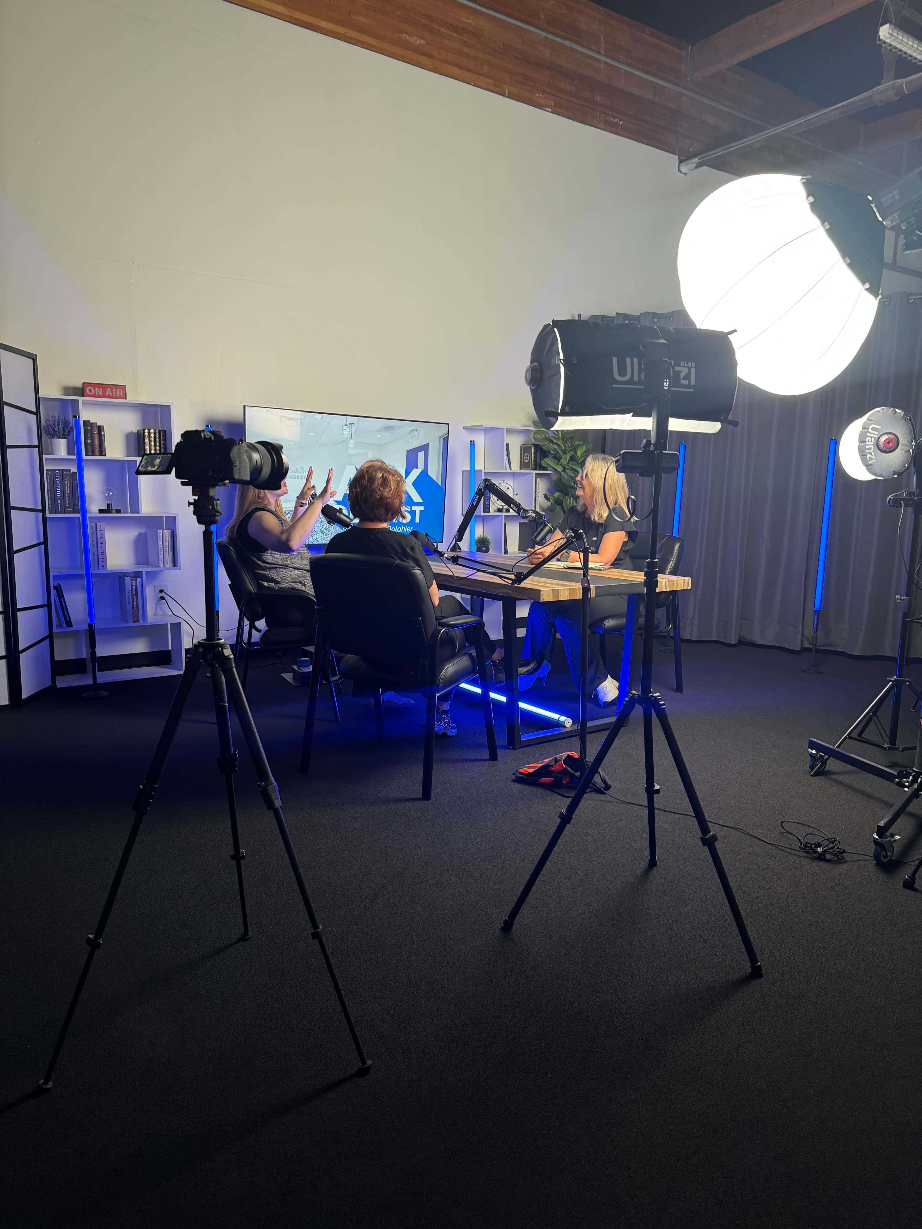 Two women are seated at a table in a studio setting, discussing while a camera and lights are set up for recording.