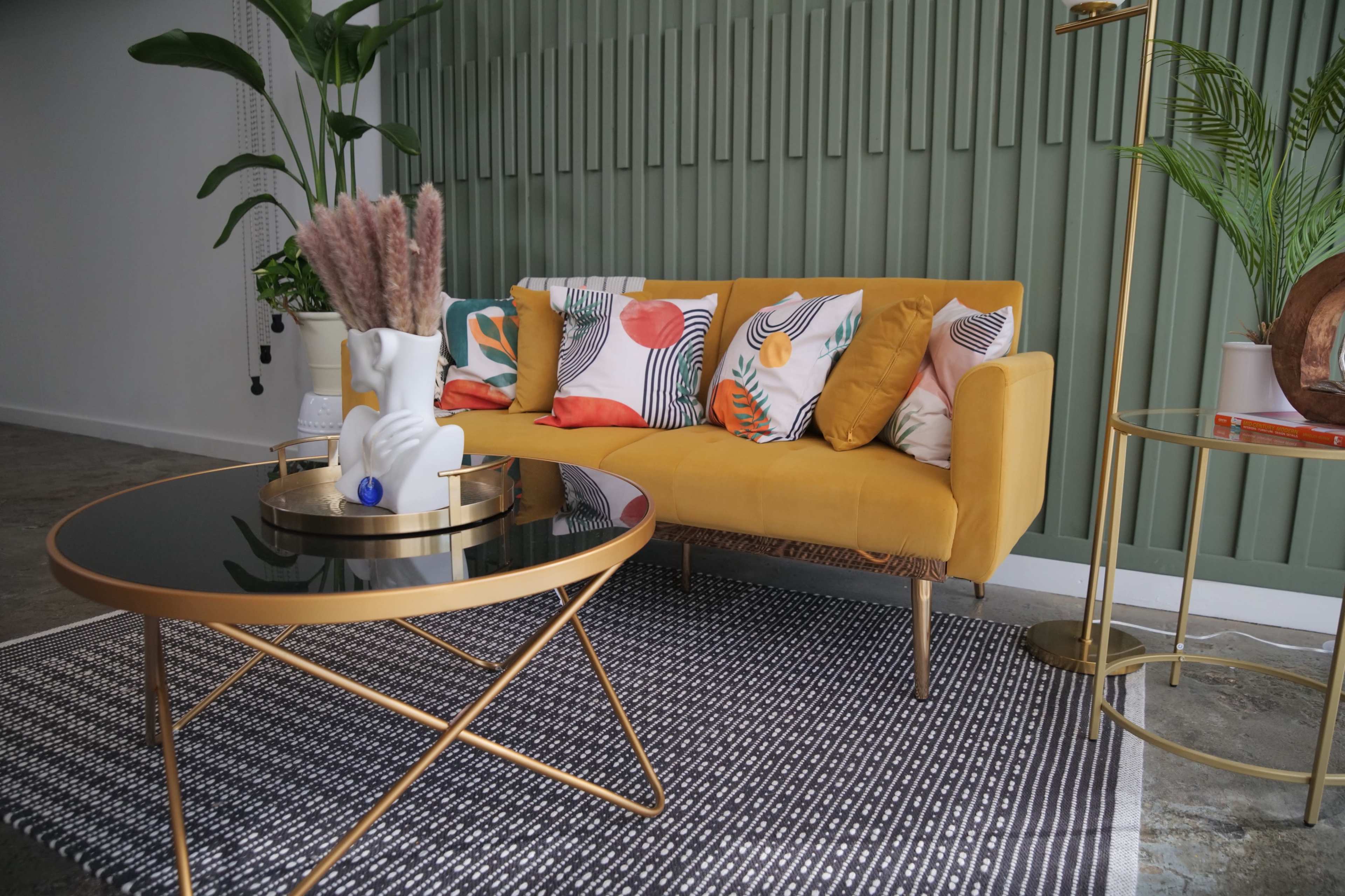 A yellow sofa with patterned cushions is positioned in a room featuring a green accent wall, a round coffee table, and plants.