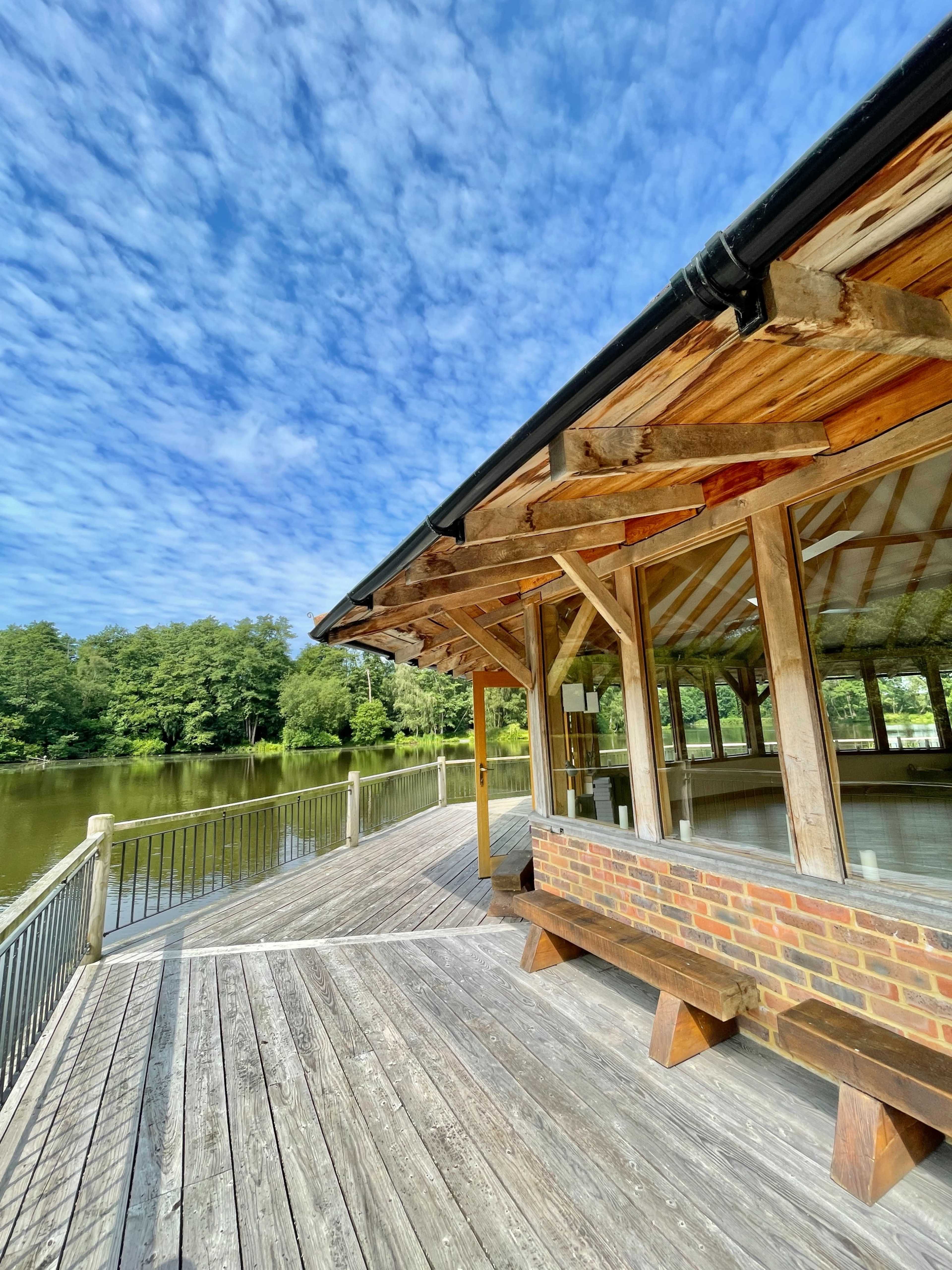 A wooden pavilion with a spacious deck overlooks a serene lake surrounded by trees under a cloudy sky.