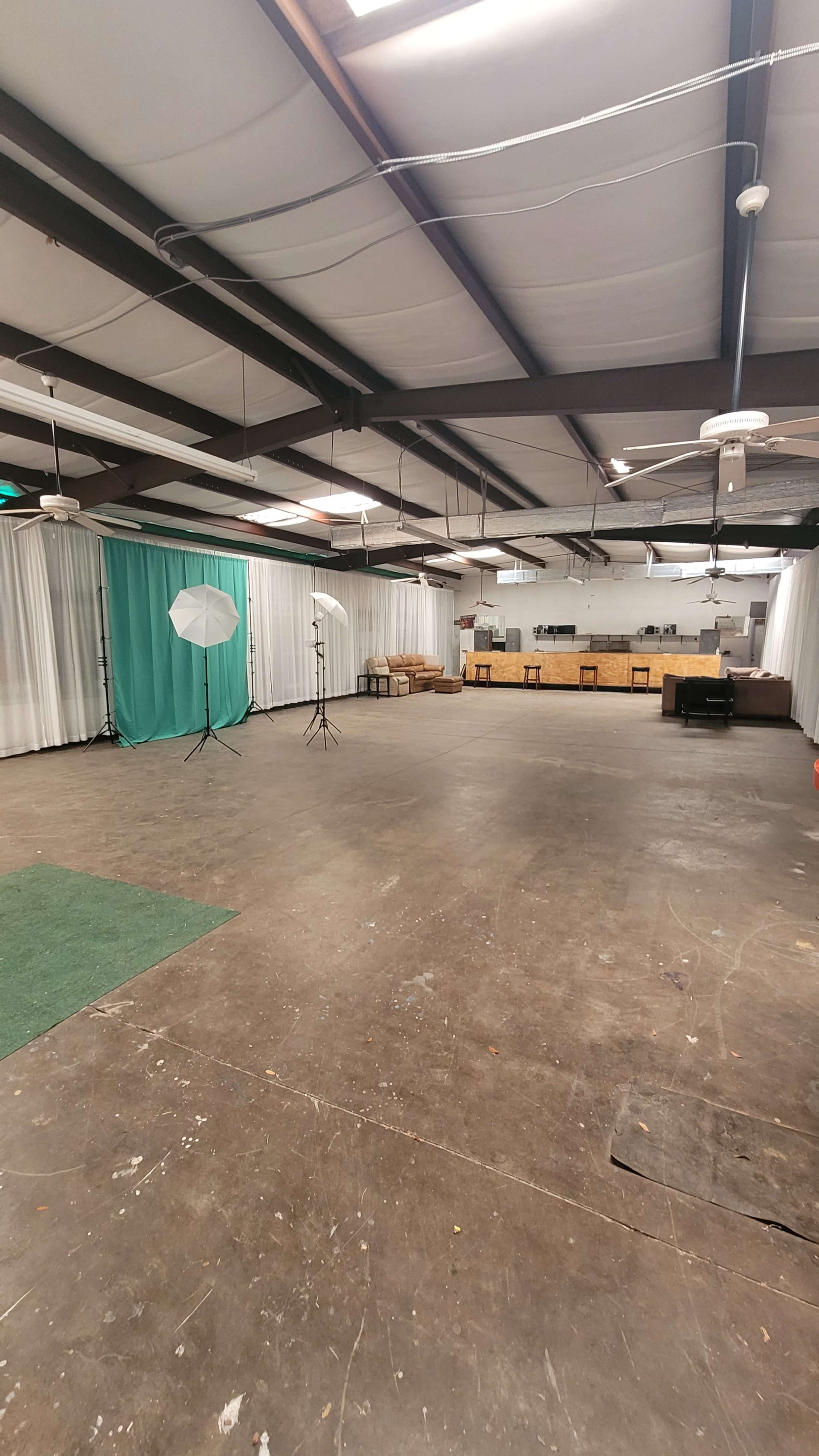 The image shows an empty indoor space with a green carpet, photography lighting equipment, and a backdrop of white drapes, along with a wooden stage area in the background.