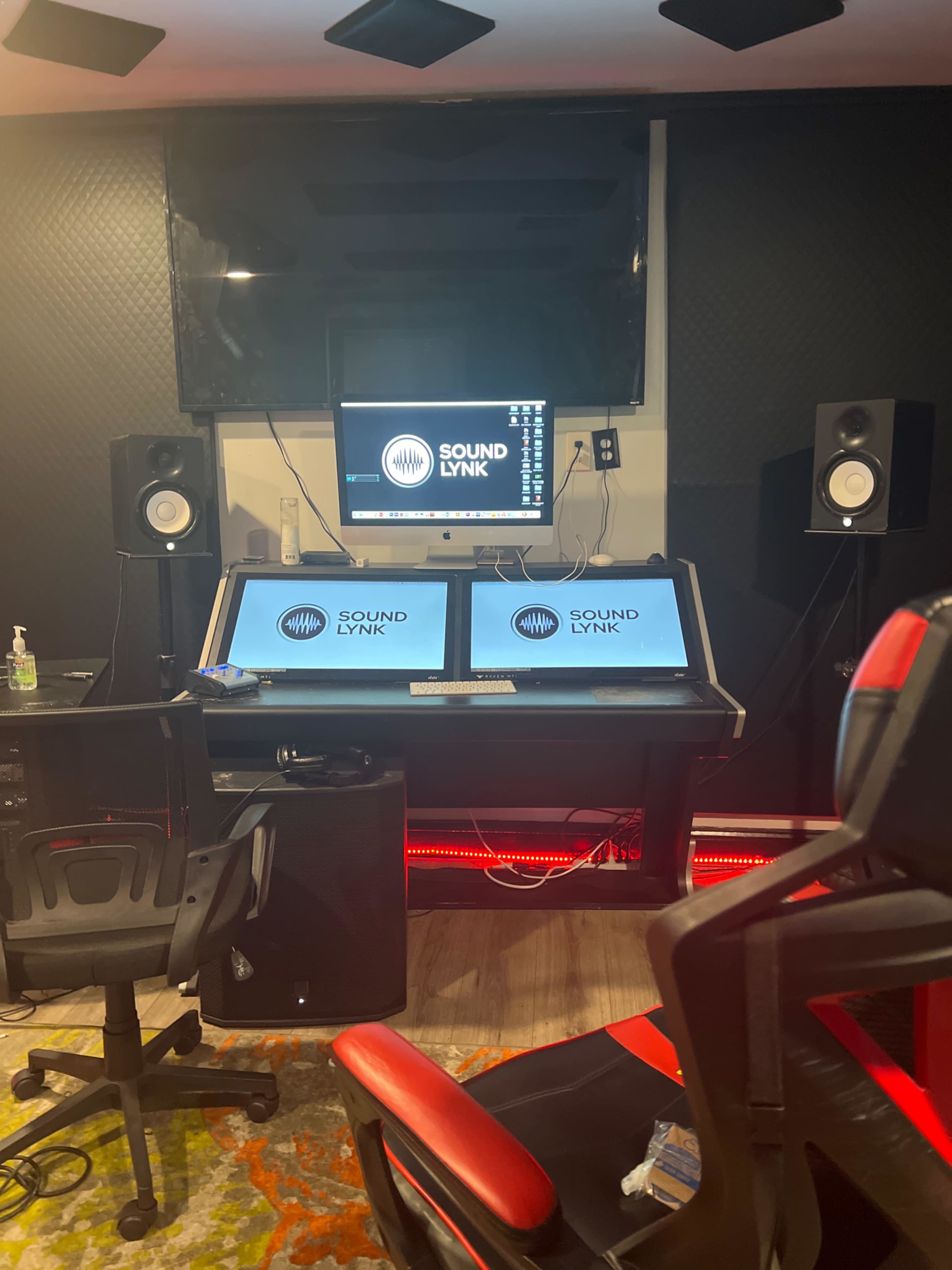 The image shows a music production studio featuring a workstation with dual monitors, studio speakers, and a red and black ergonomic chair.
