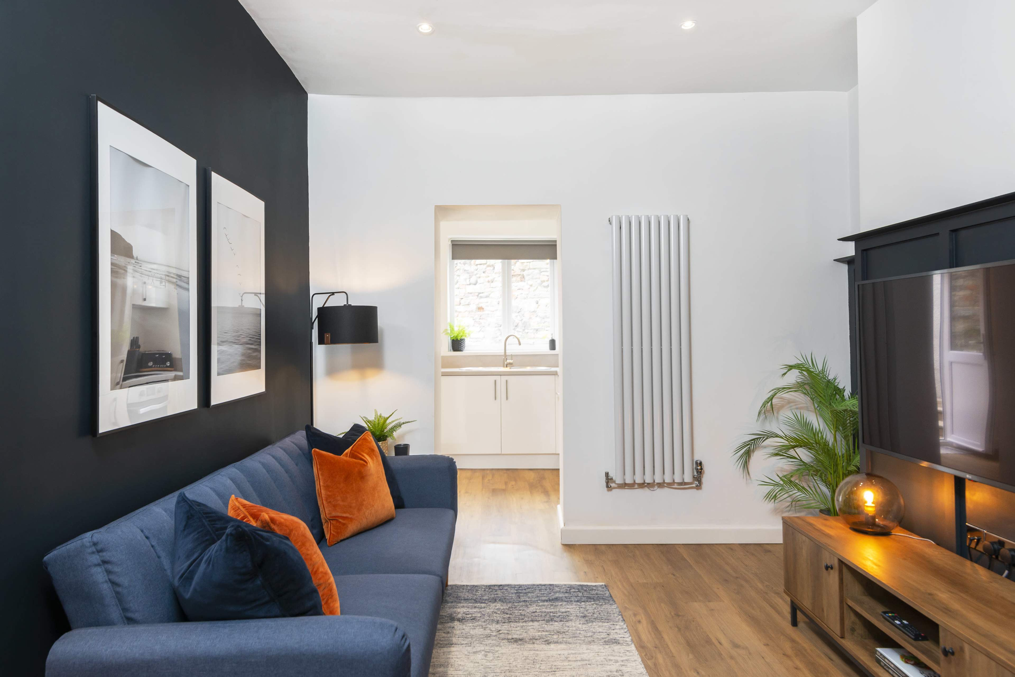 A minimalist living room with a dark accent wall, a blue sofa adorned with orange and blue cushions, and a window leading to a kitchen area.