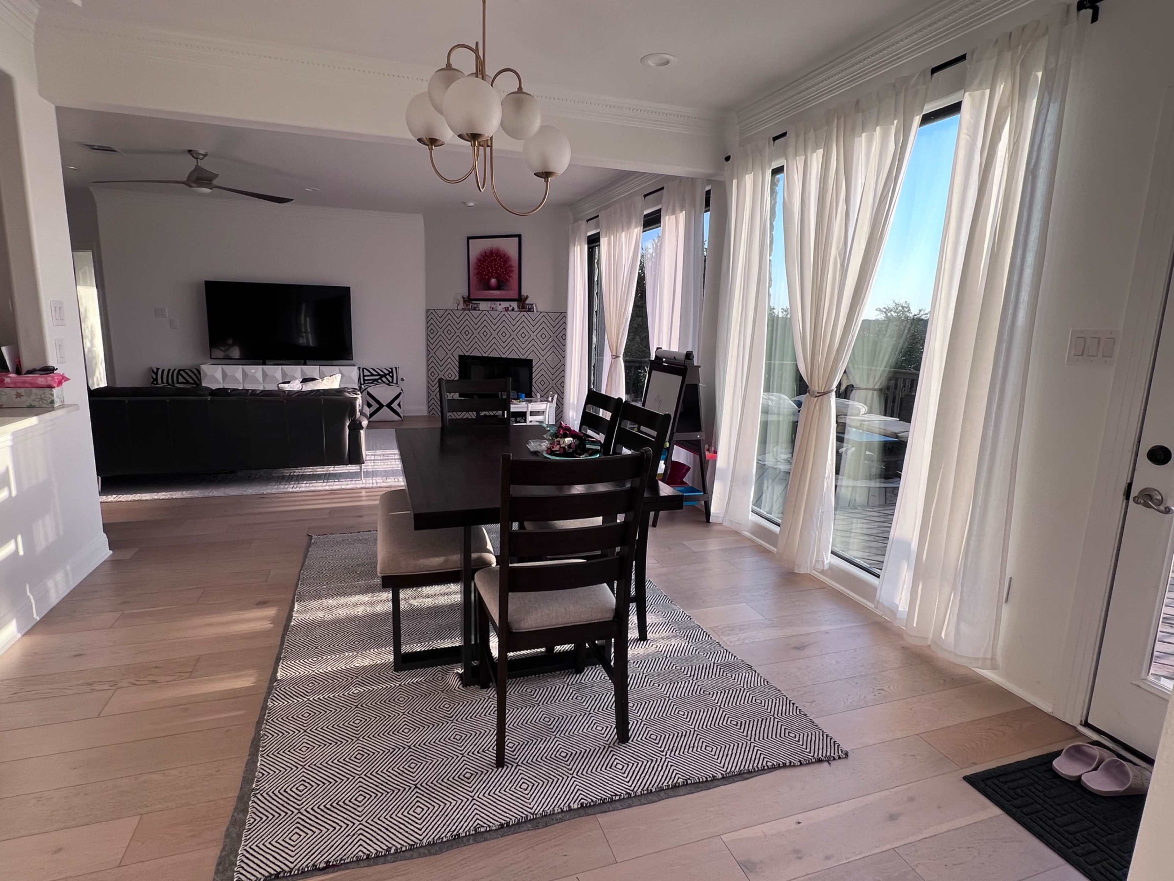 The image shows a modern dining area with a dark wooden table and chairs, surrounded by large windows with sheer curtains, leading into a living space featuring a television and a fireplace.