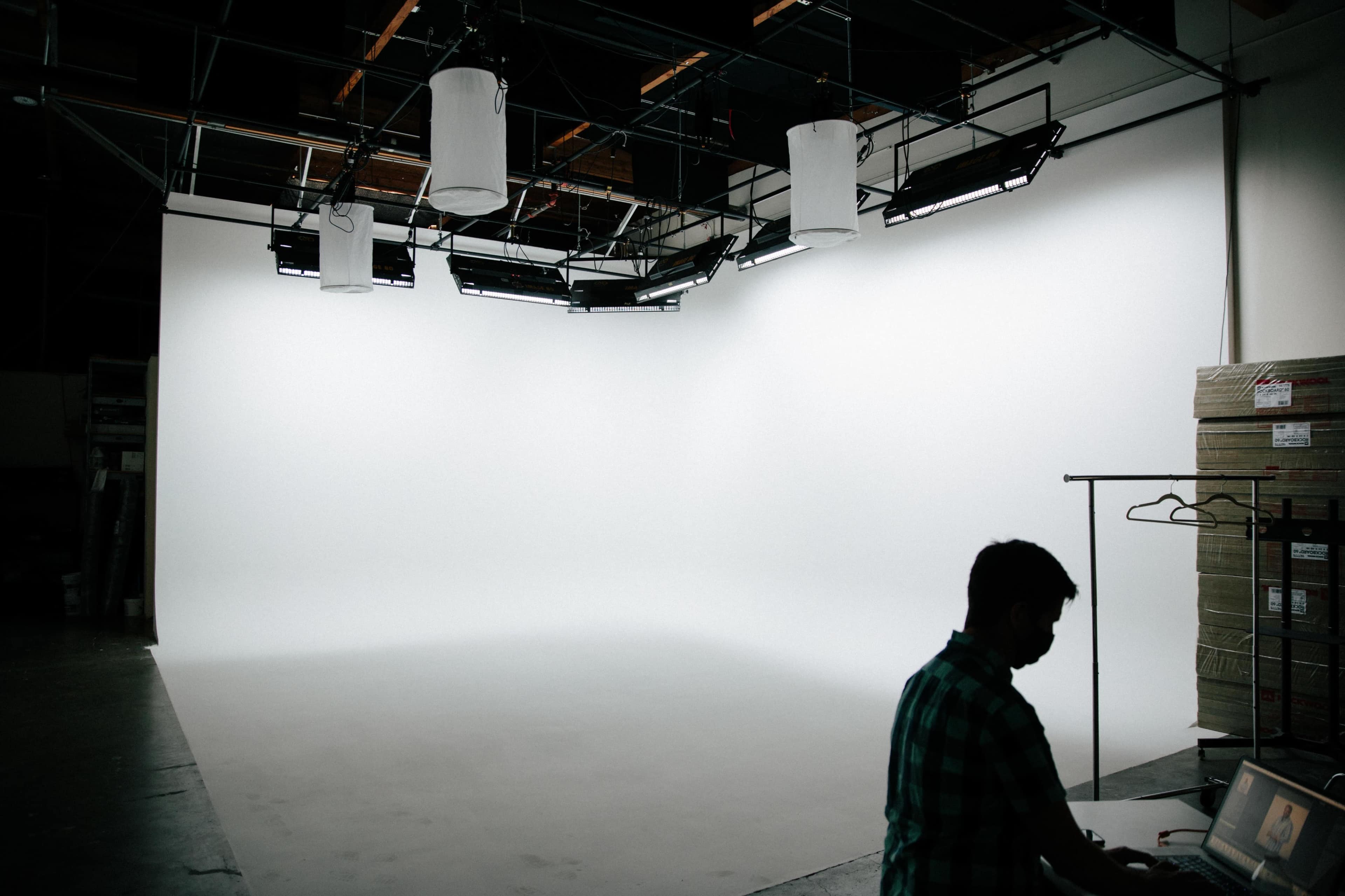 A person works at a table in a spacious, well-lit studio with a seamless white backdrop and overhead lights.