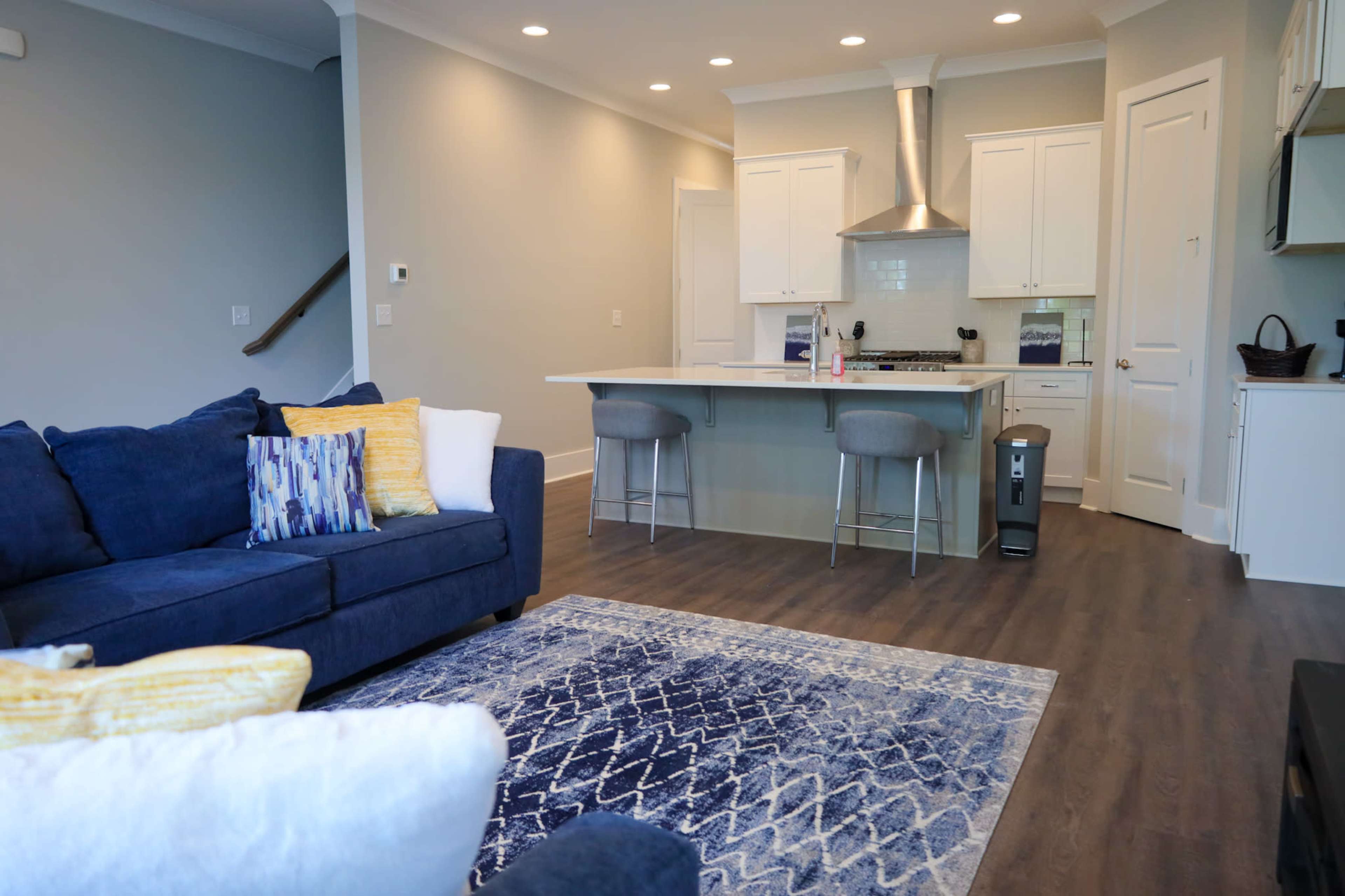 The image shows a modern living room with a blue sectional sofa, a light-colored kitchen in the background featuring a central island and stainless steel appliances, and a patterned area rug on the floor.